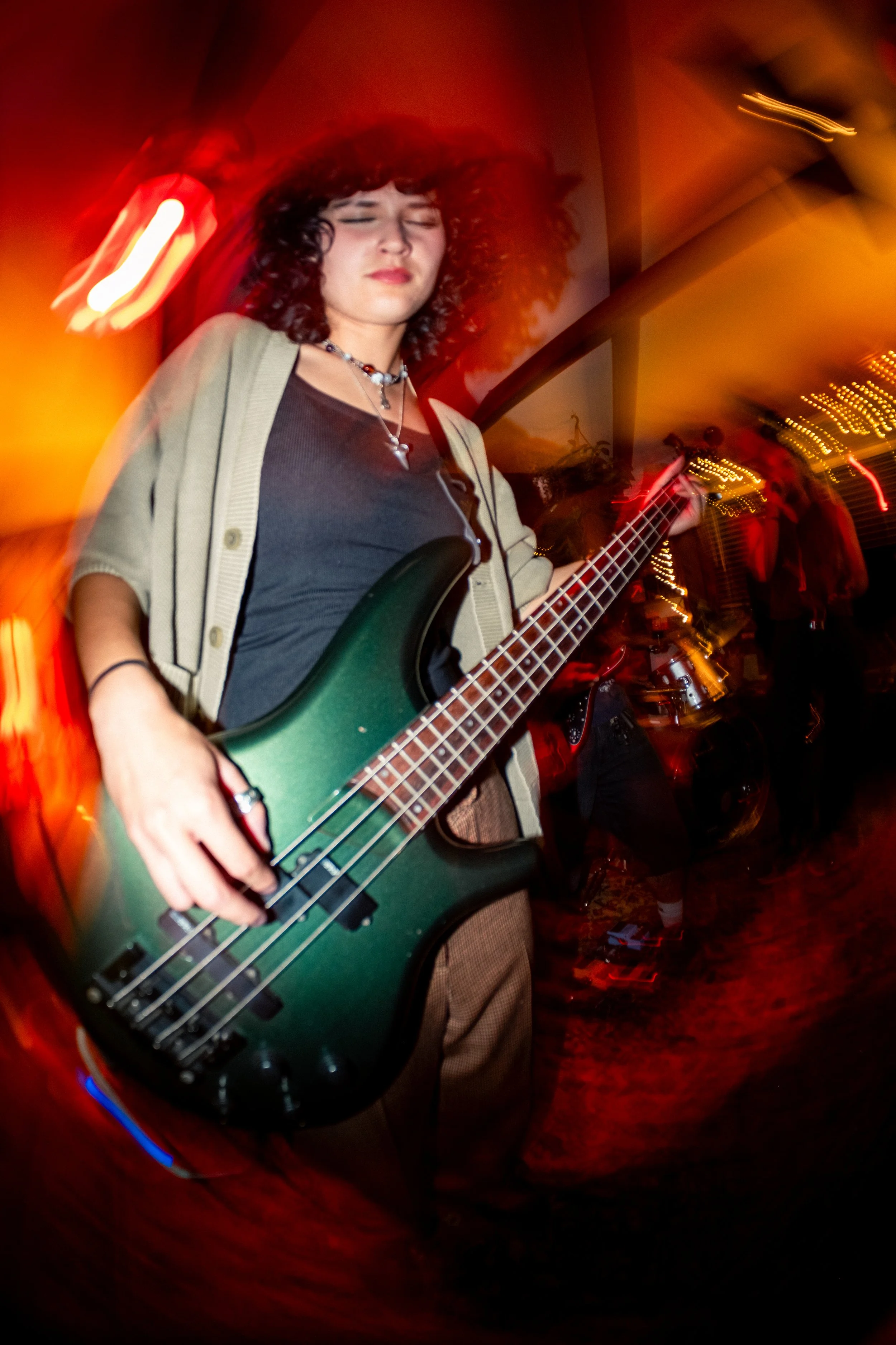 A woman with curly hair playing an electric bass guitar with her eyes closed, in a dimly lit room with warm lighting and people in the background.