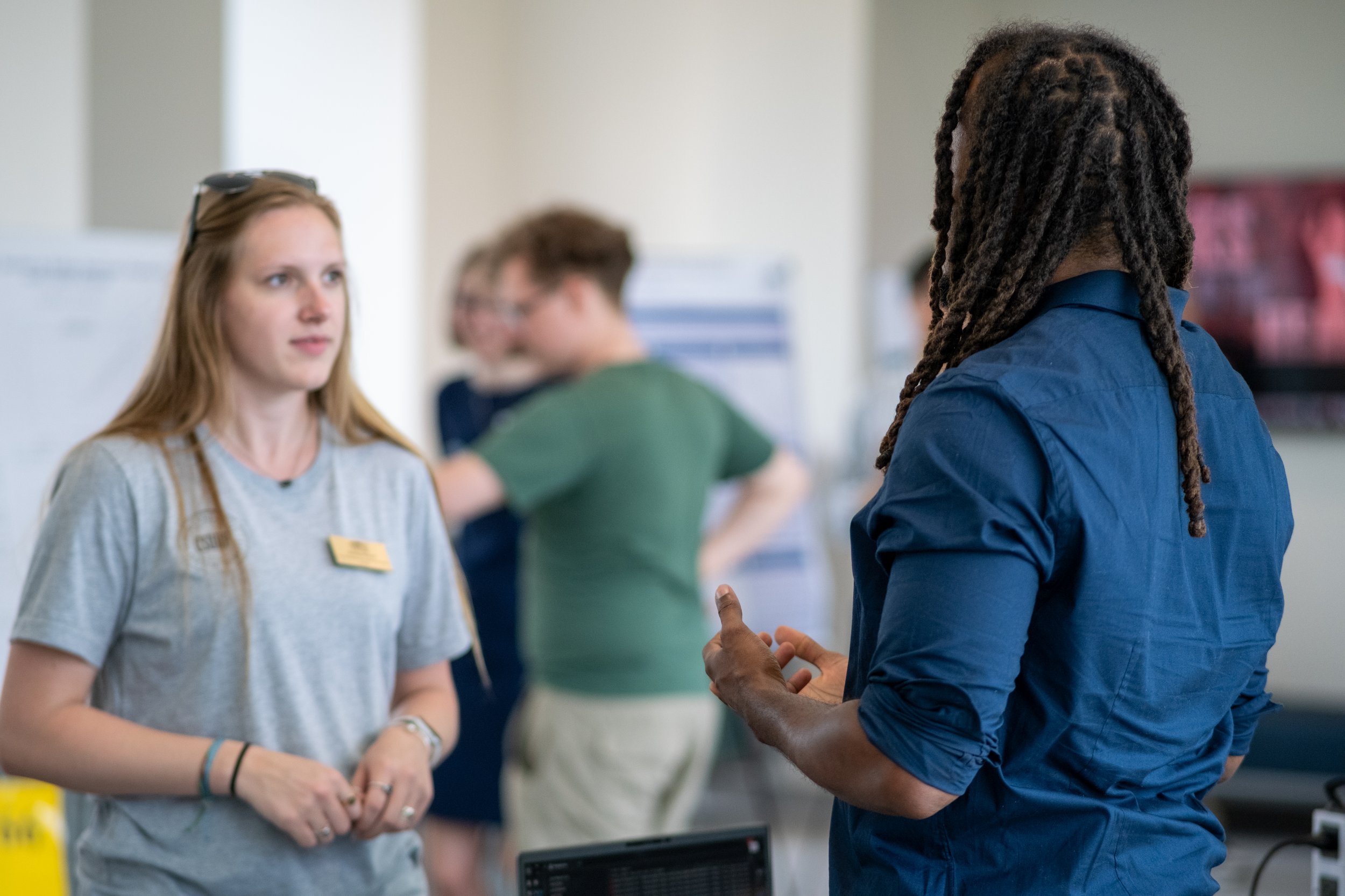 A woman with blonde hair and a man with dark skin and dreadlocks are having a conversation in a public indoor space. The woman is wearing a light gray t-shirt and a name tag, while the man is wearing a blue shirt. There are other people in the backgr