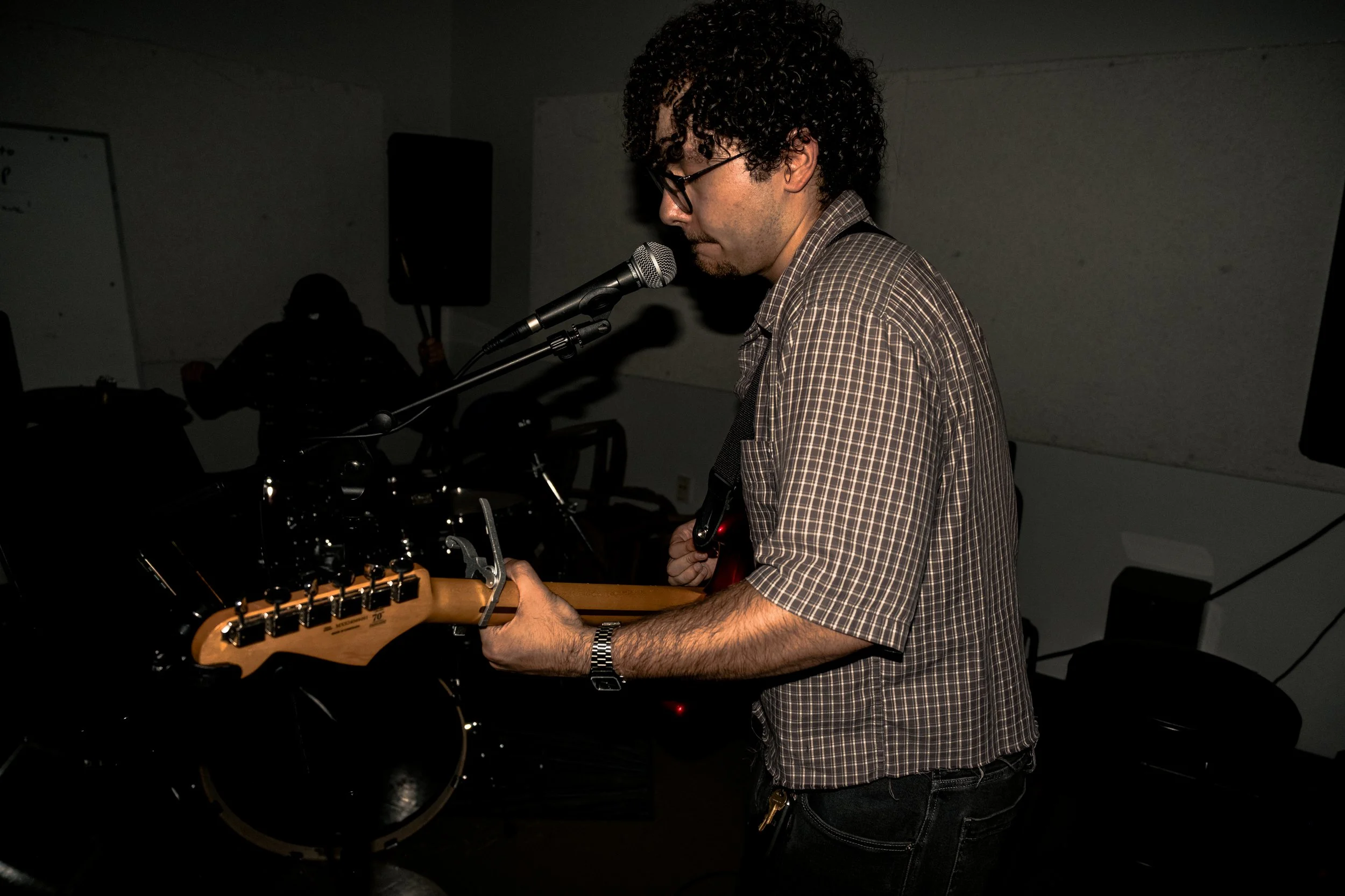 A man with curly hair, glasses, wearing a checkered shirt, playing an electric guitar and singing into a microphone in a dimly lit room.