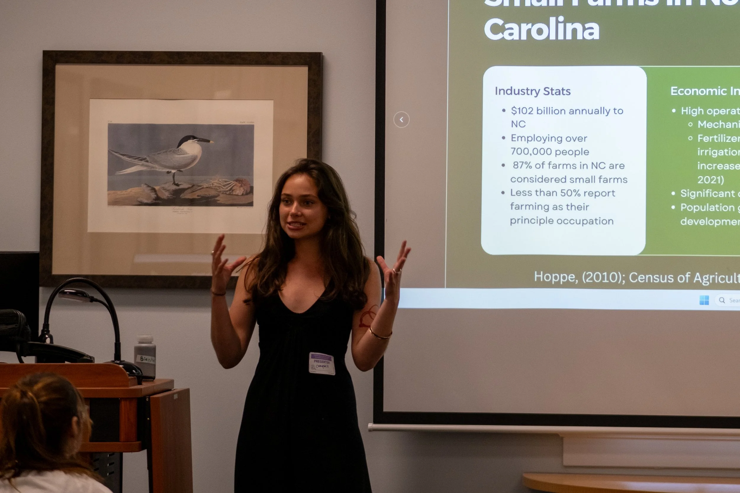 A woman giving a presentation in a classroom or conference room. She is standing in front of a large screen displaying a slide with text about industry statistics and economic information. She has long hair, is wearing a sleeveless top, and is gestur