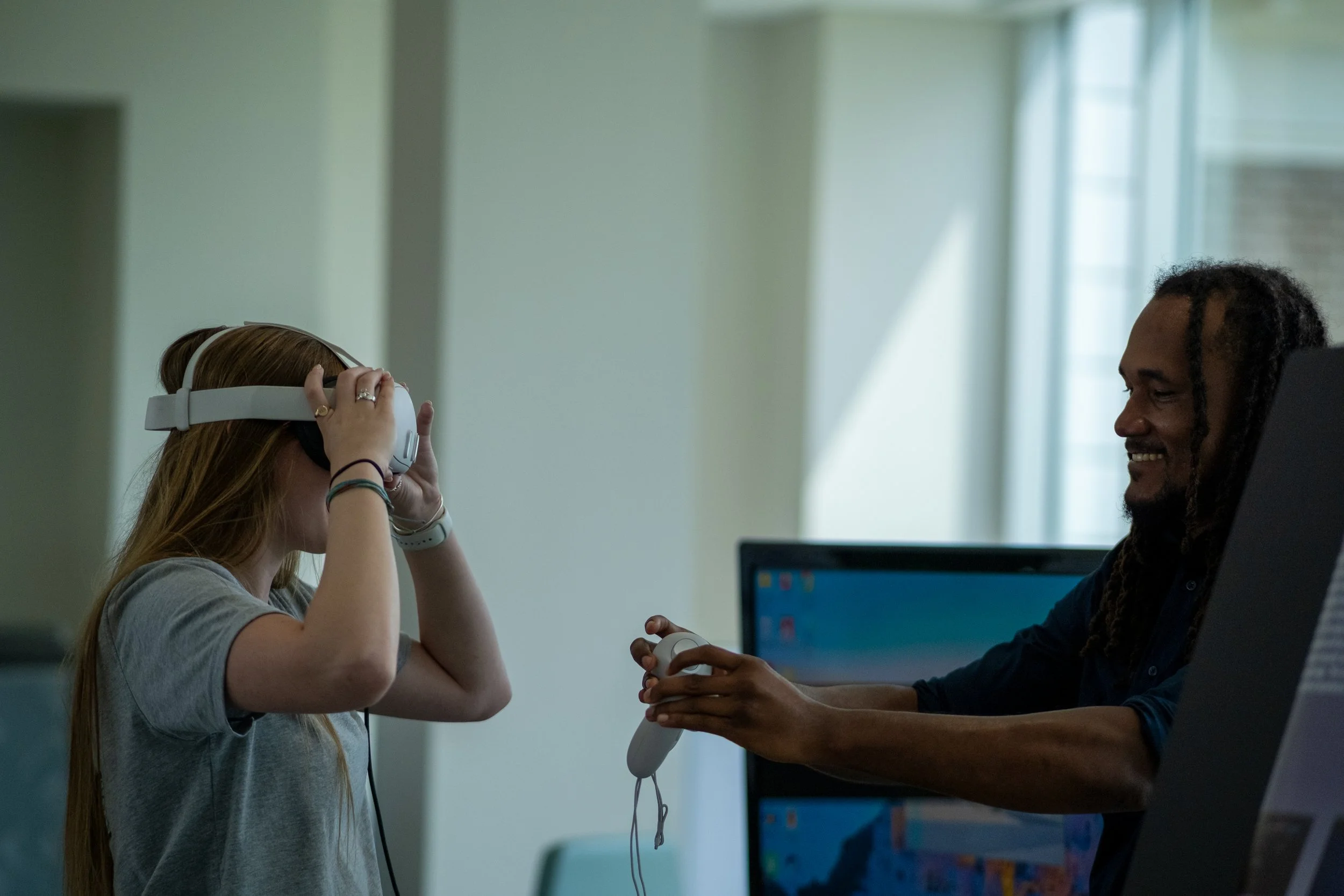 A woman with long red hair wearing headphones and a gray shirt is adjusting her headphones while sitting at a desk. A man with long braided hair, smiling, is holding an electronic device and sitting across from her. There is a computer monitor with a