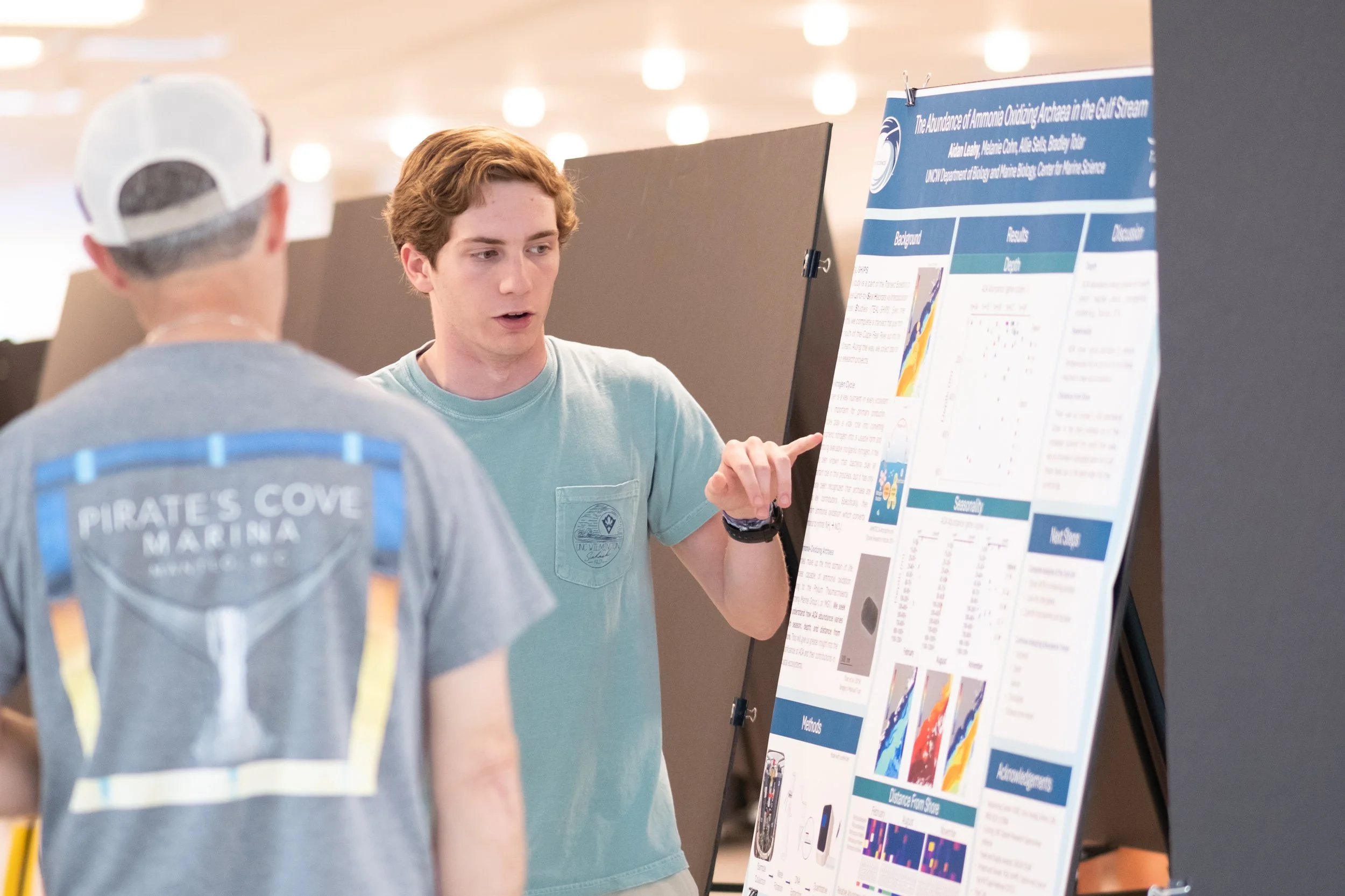 Two young men having a discussion in front of a scientific research poster at an academic conference. The poster discusses the abundance of ammonia oxidizing archaea in the Gulf Stream.