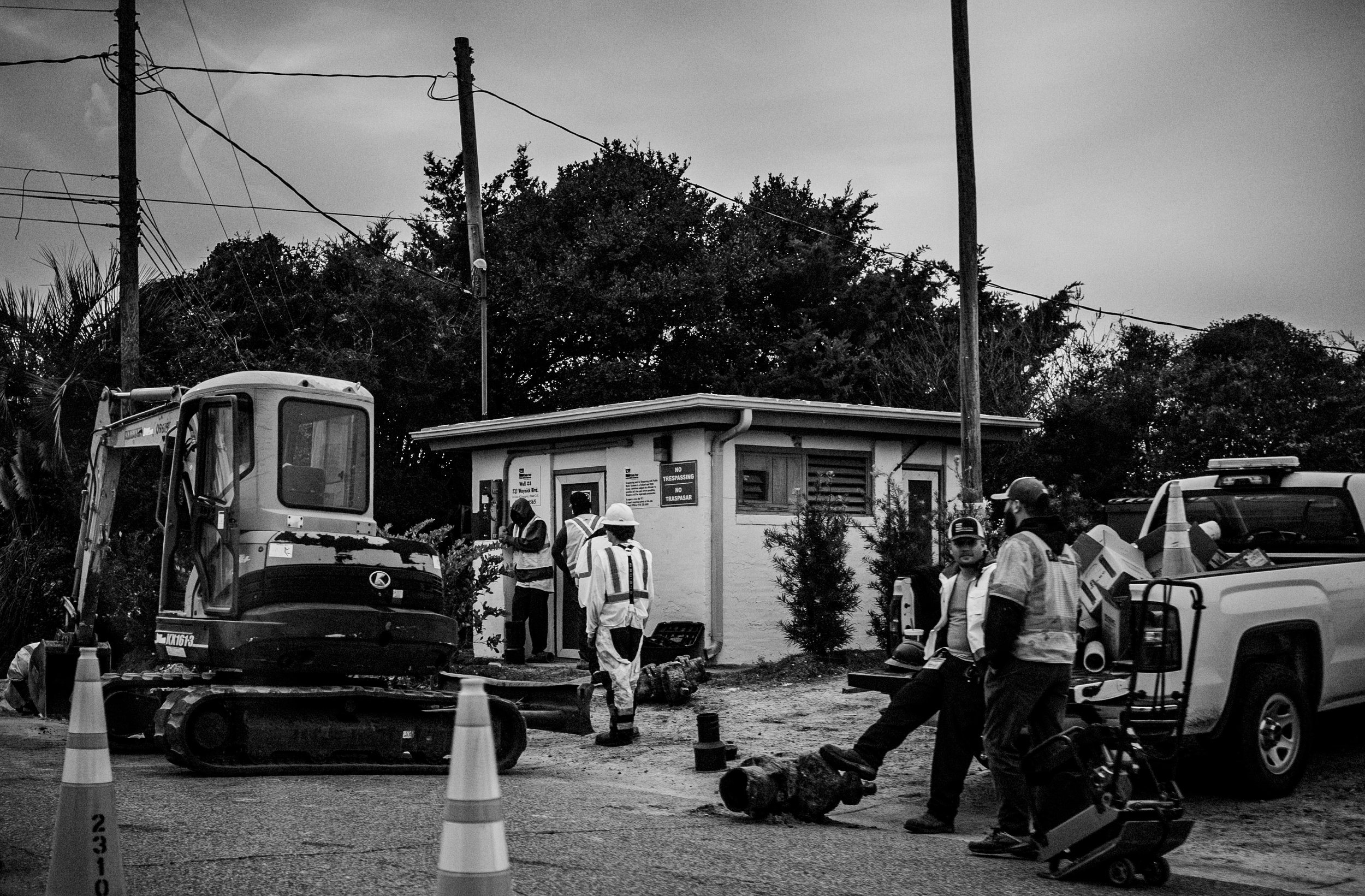 Construction workers and equipment at a construction site with a small building, trees, and utility poles in black-and-white.
