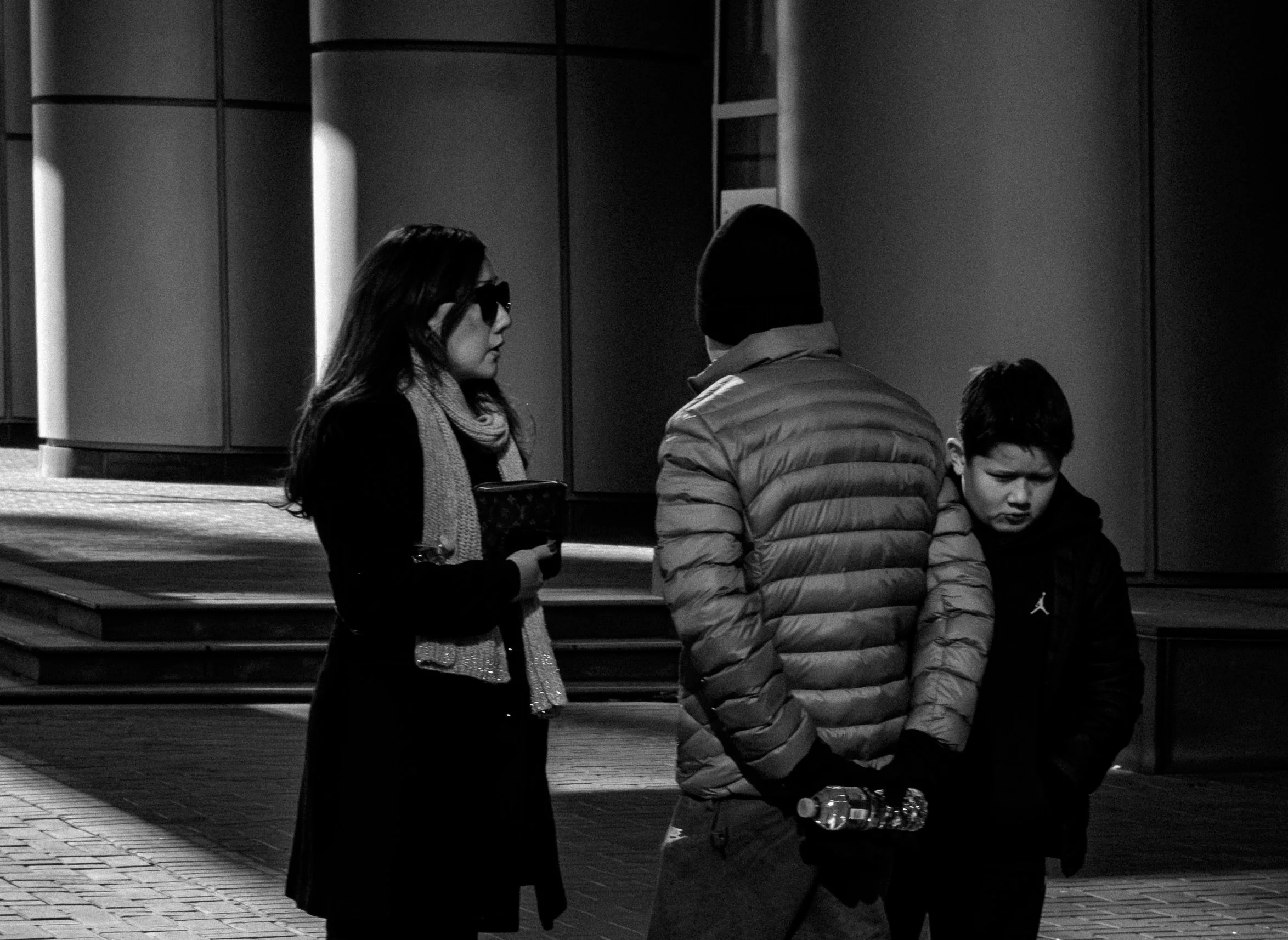 Three people, a woman and two boys, standing outdoors at night having a conversation. The woman is wearing sunglasses and a scarf, holding an item in her hand. One boy is wearing a puffer jacket with a hood, holding a water bottle, and the other boy 