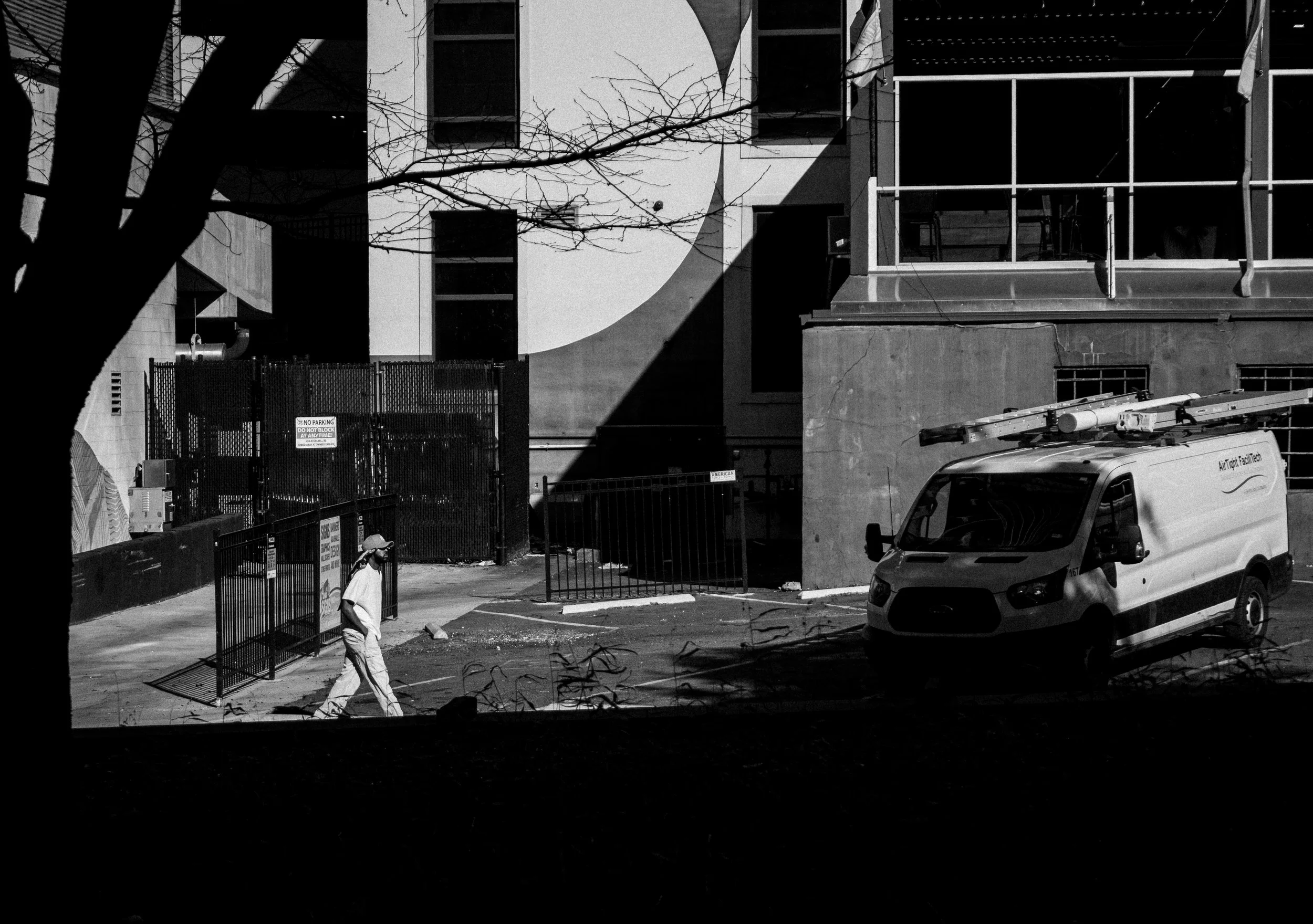 A black and white photo of an urban street scene showing a person walking past a gated alley, a white service van with ladders on top, and a modern building with a balcony and large windows. Leafless tree branches frame the scene.