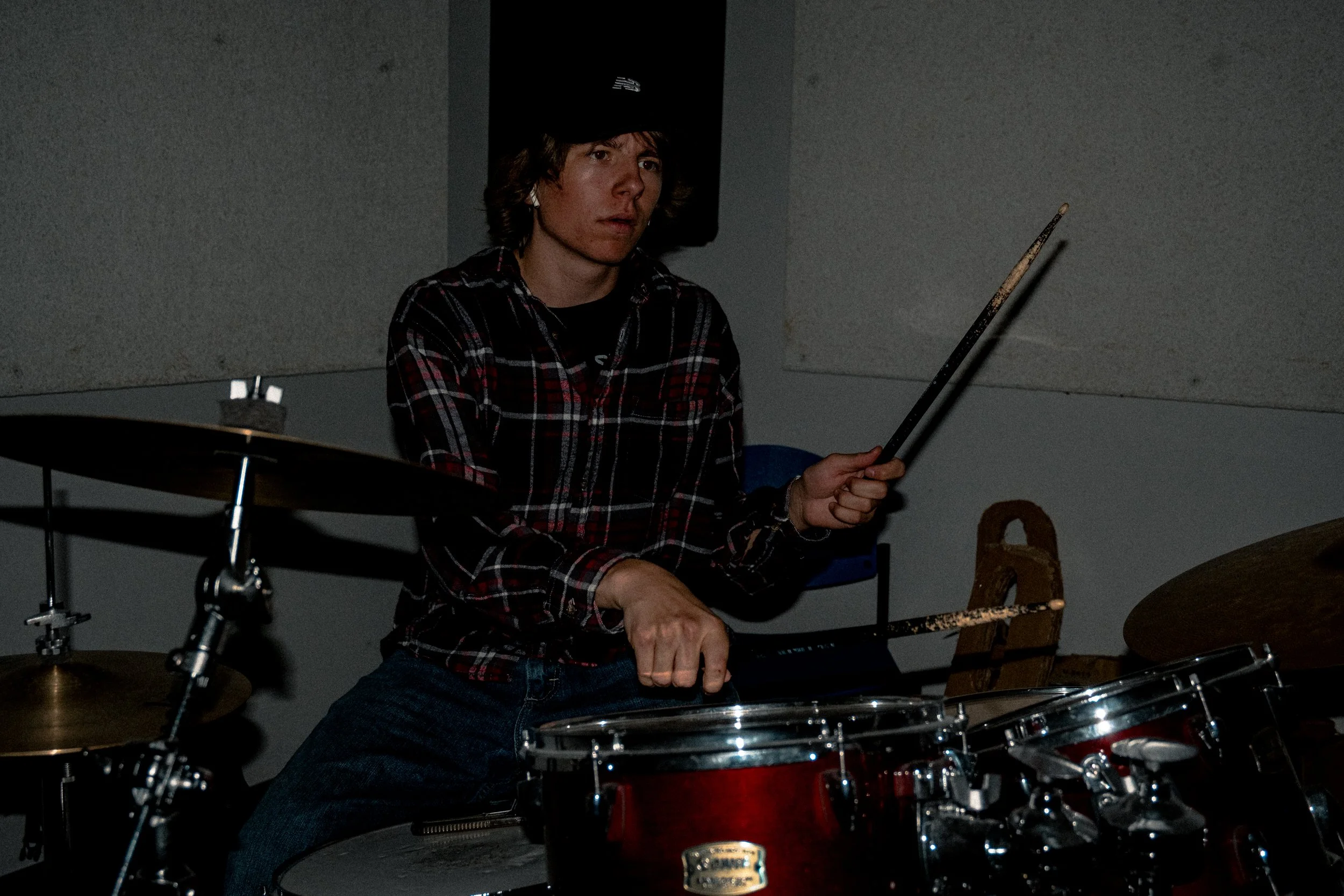 A young man with brown hair playing a drum set, wearing a black cap and a red plaid shirt in a dimly lit room.