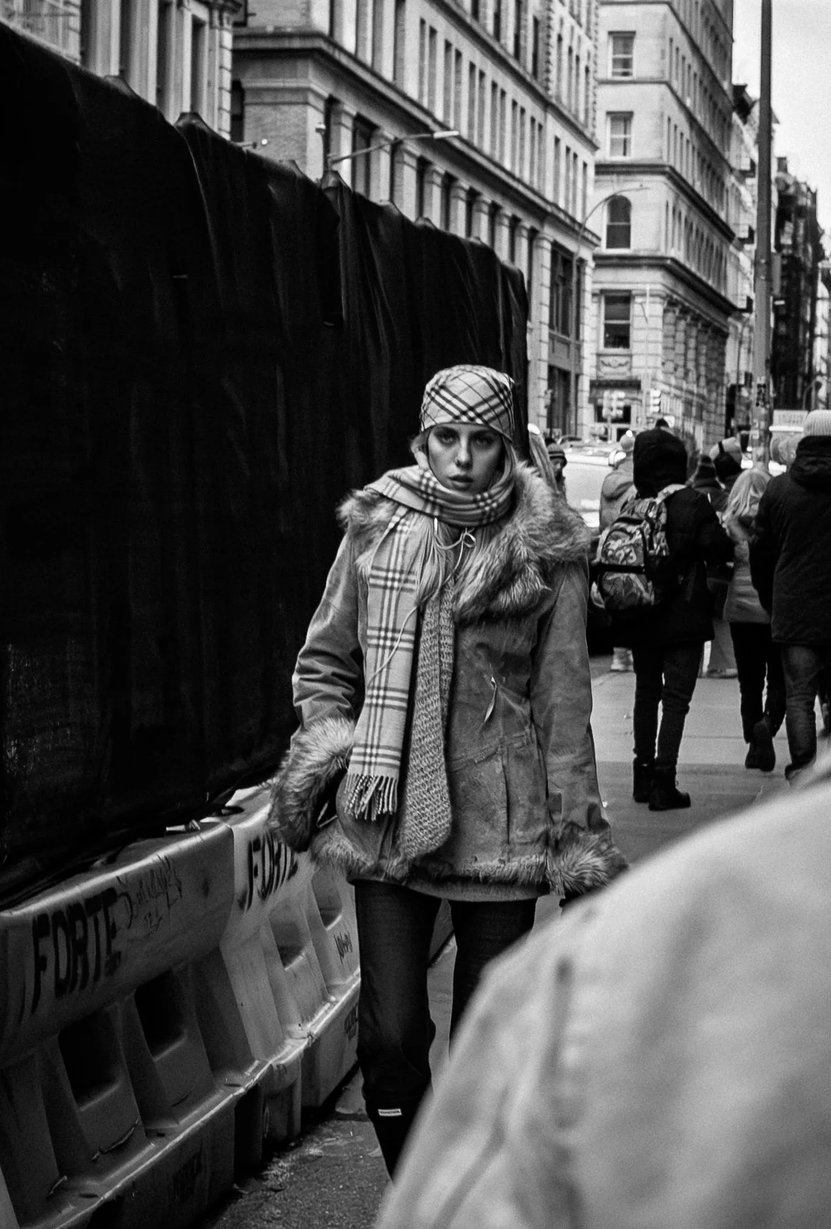 A woman walking on a city street in winter, wearing a fur-lined coat, plaid scarf, and matching plaid hat, with tall buildings and other pedestrians in the background.