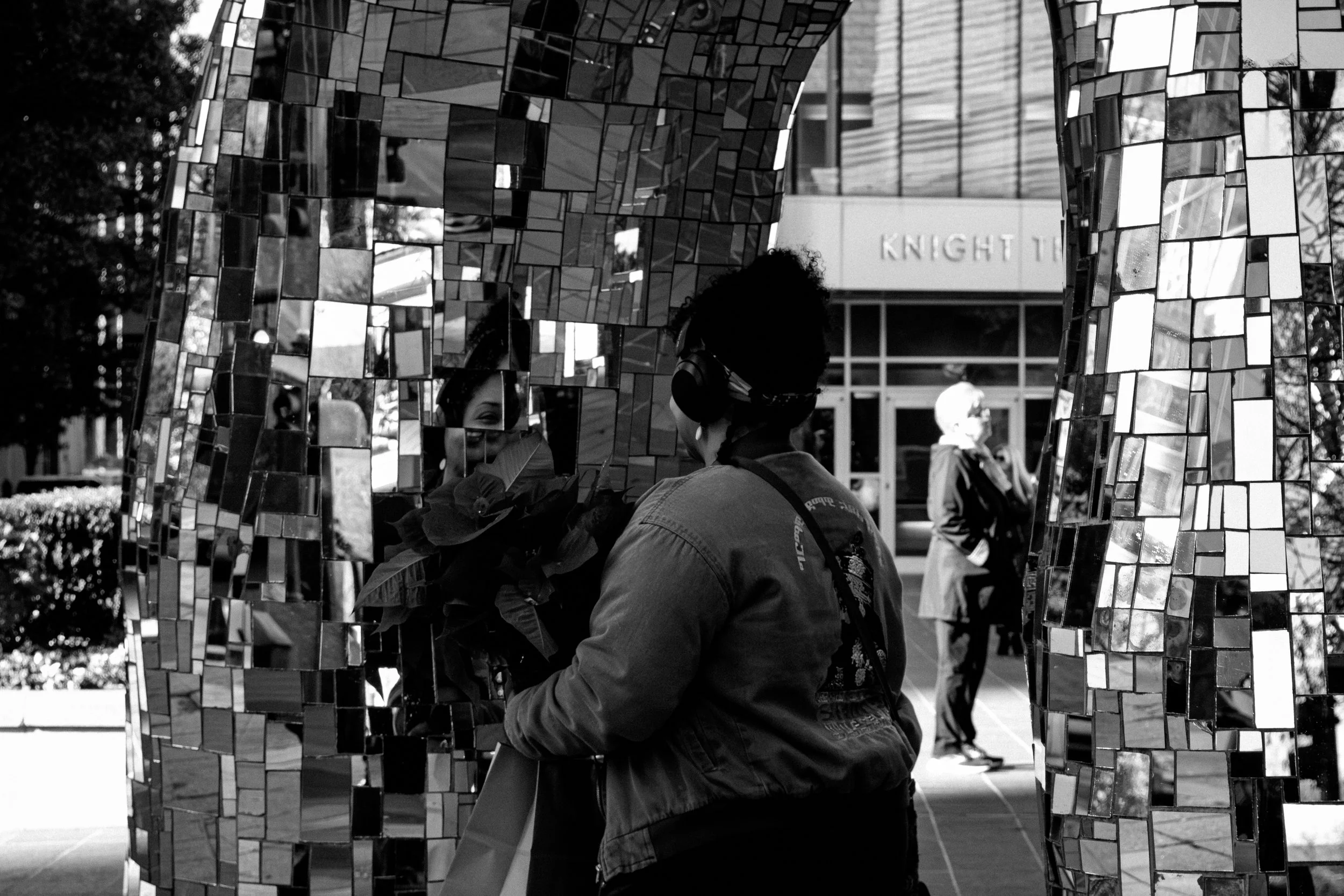 A woman looking at her reflection in a mirror mosaic sculpture outside a building titled 'KNIGHT' in the background.