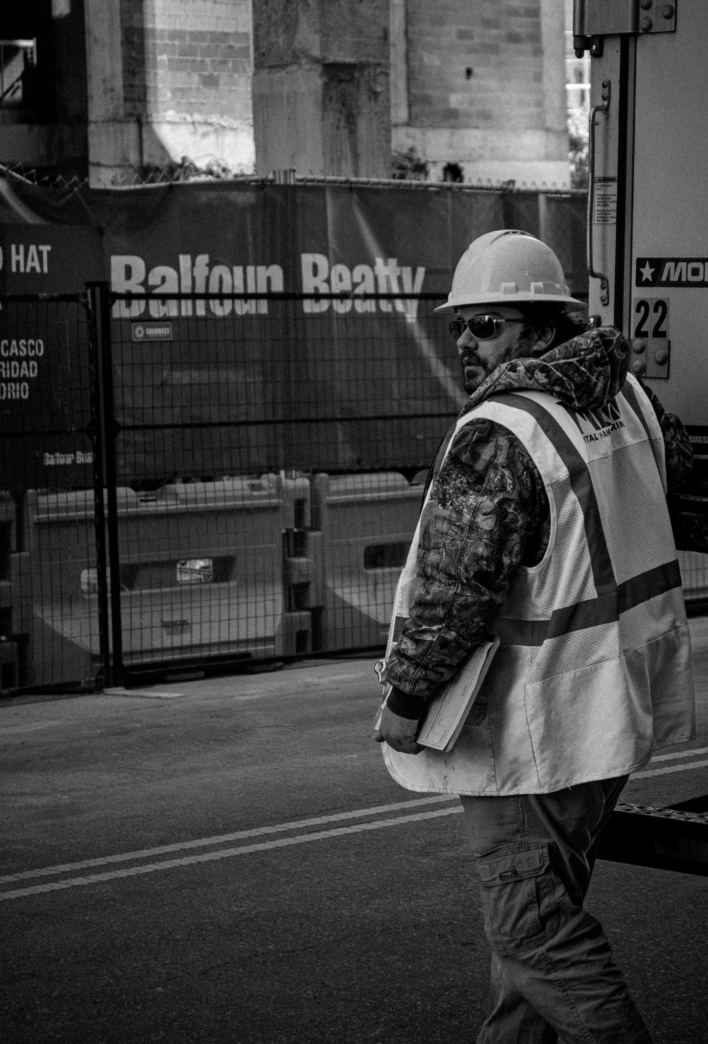 A construction worker wearing a hard hat, safety glasses, and a reflective vest stands on the street holding a clipboard, with a construction site and barriers in the background.
