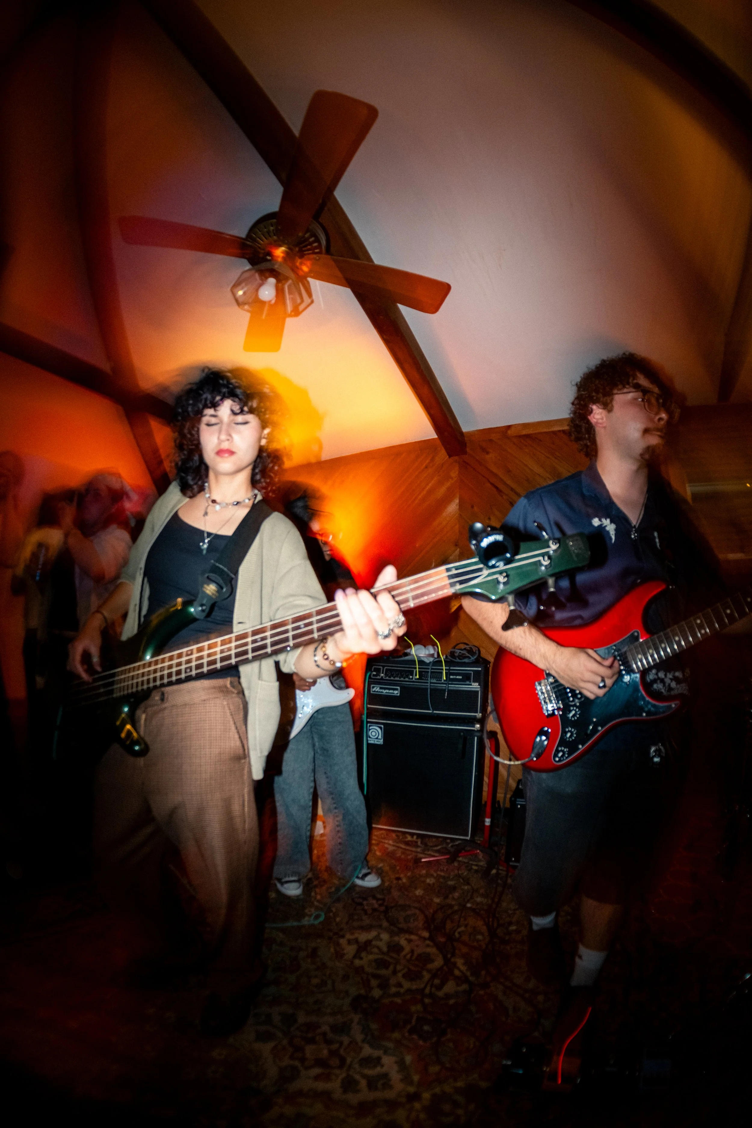Two musicians playing electric guitars at a live music event in a cozy, wood-paneled room with a ceiling fan. The woman on the left has curly hair, dark lipstick, and wears a beige cardigan over a black top. The man on the right has curly hair, glass