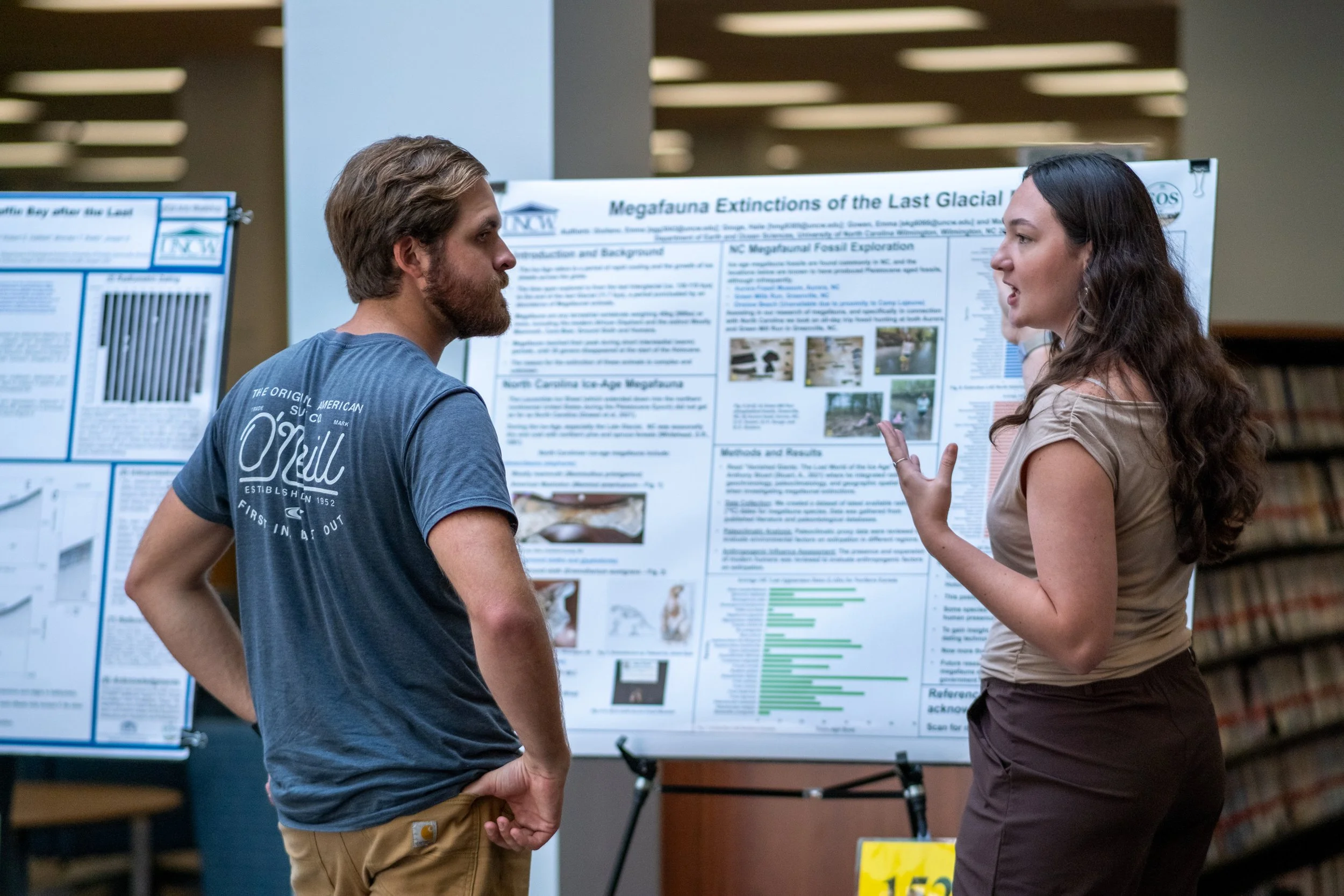 A man and woman are engaged in conversation at a research poster presentation. The man has light brown hair and a beard, wearing a blue t-shirt with white text. The woman has long dark hair and is wearing a beige top with earrings, gesturing as she e