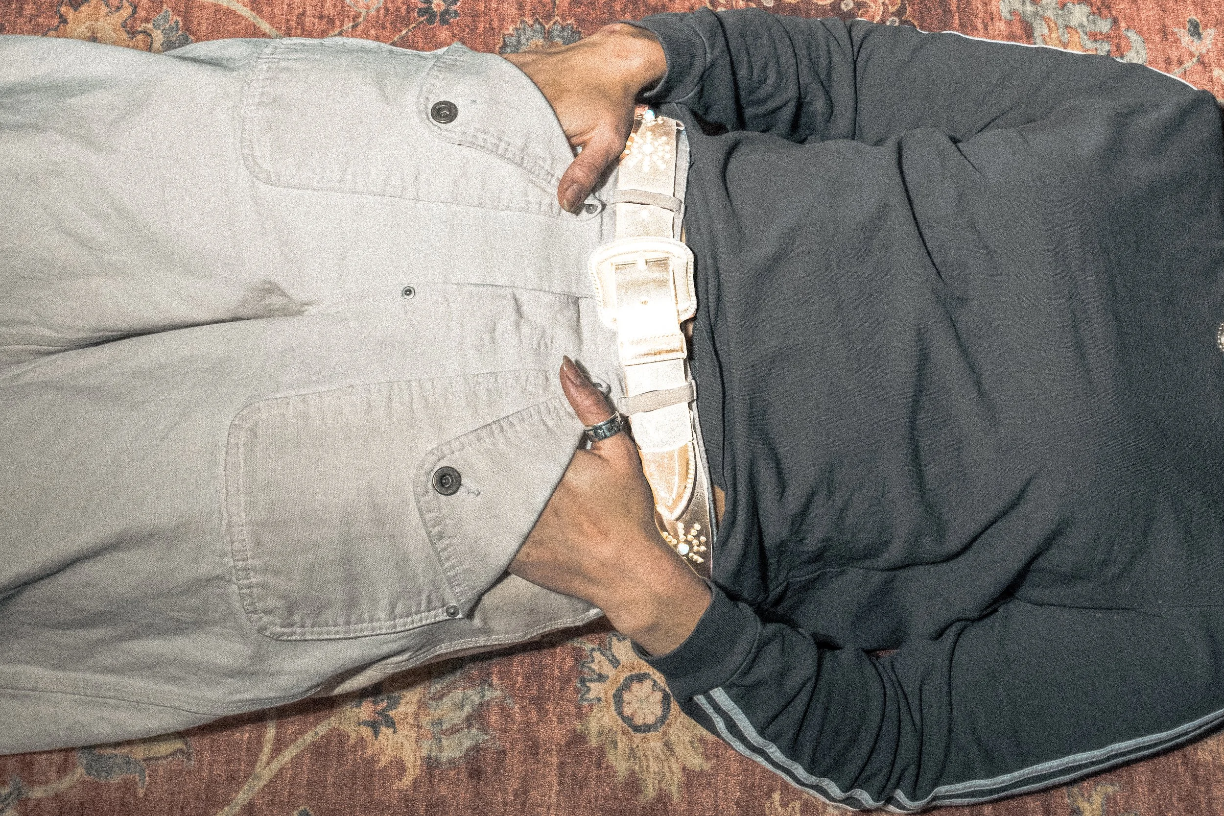 A person lying on their side on a patterned rug, wearing gray pants, a black jacket, and a white shirt. Their hand is resting on their hip, and they are holding a lit joint.