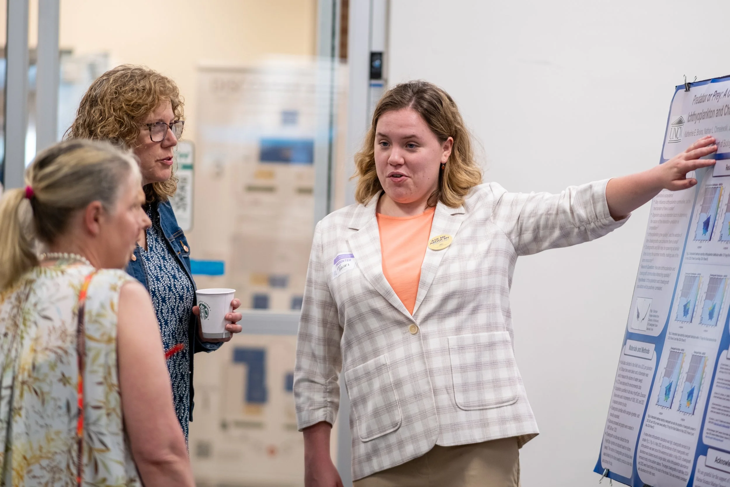 A woman with a name tag pointing to a science research poster at a conference, speaking with two women, one with glasses and curly hair, and the other with blonde hair tied back.