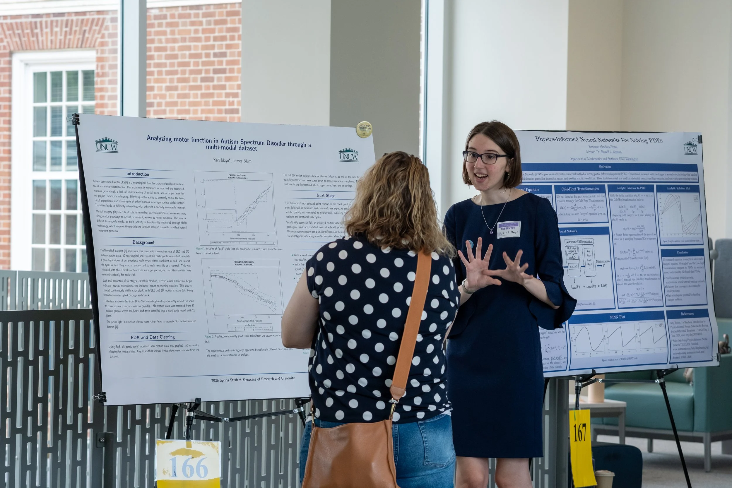 A woman with short brown hair and glasses, wearing a navy blazer and a necklace, is talking to another woman with long blonde hair wearing a black and white polka dot shirt at a research poster presentation. The research poster behind her discusses a