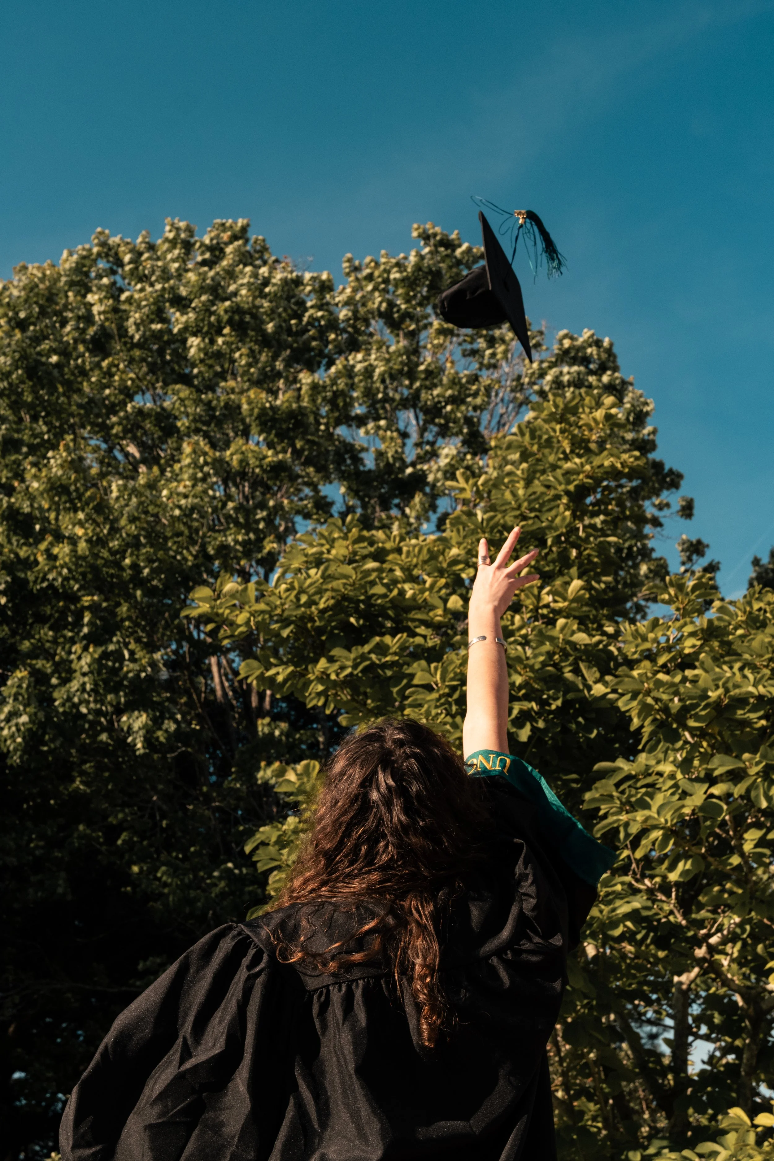 A person with long curly hair wearing a black gown and raising their hand throws a graduation cap into the air outdoors with green trees and blue sky in the background.