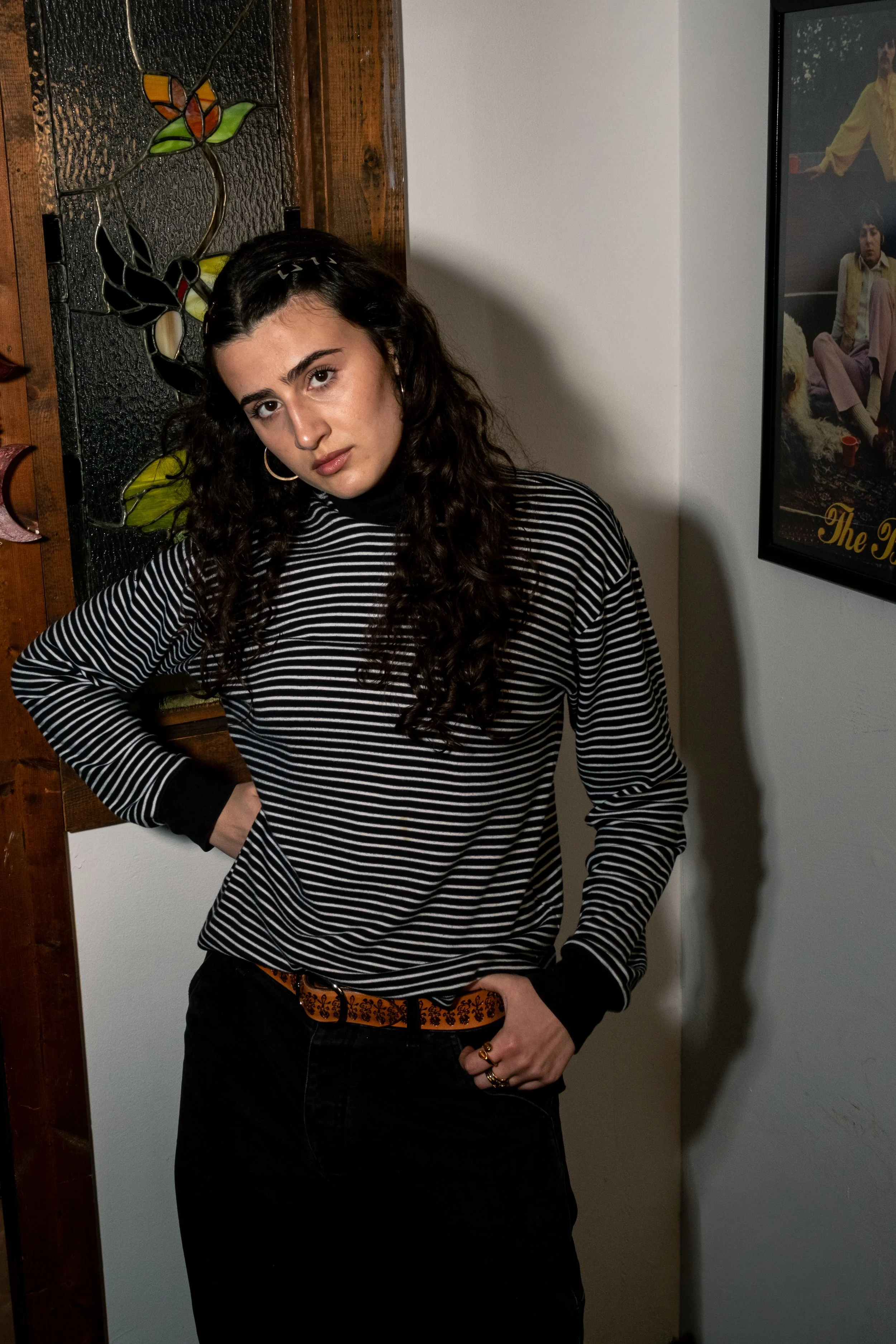 A young woman with dark curly hair, wearing a black and white striped long sleeve top and black pants, standing indoors with her left hand on her hip, looking at the camera. Behind her is a wood panel with stained glass and a framed poster on the wal