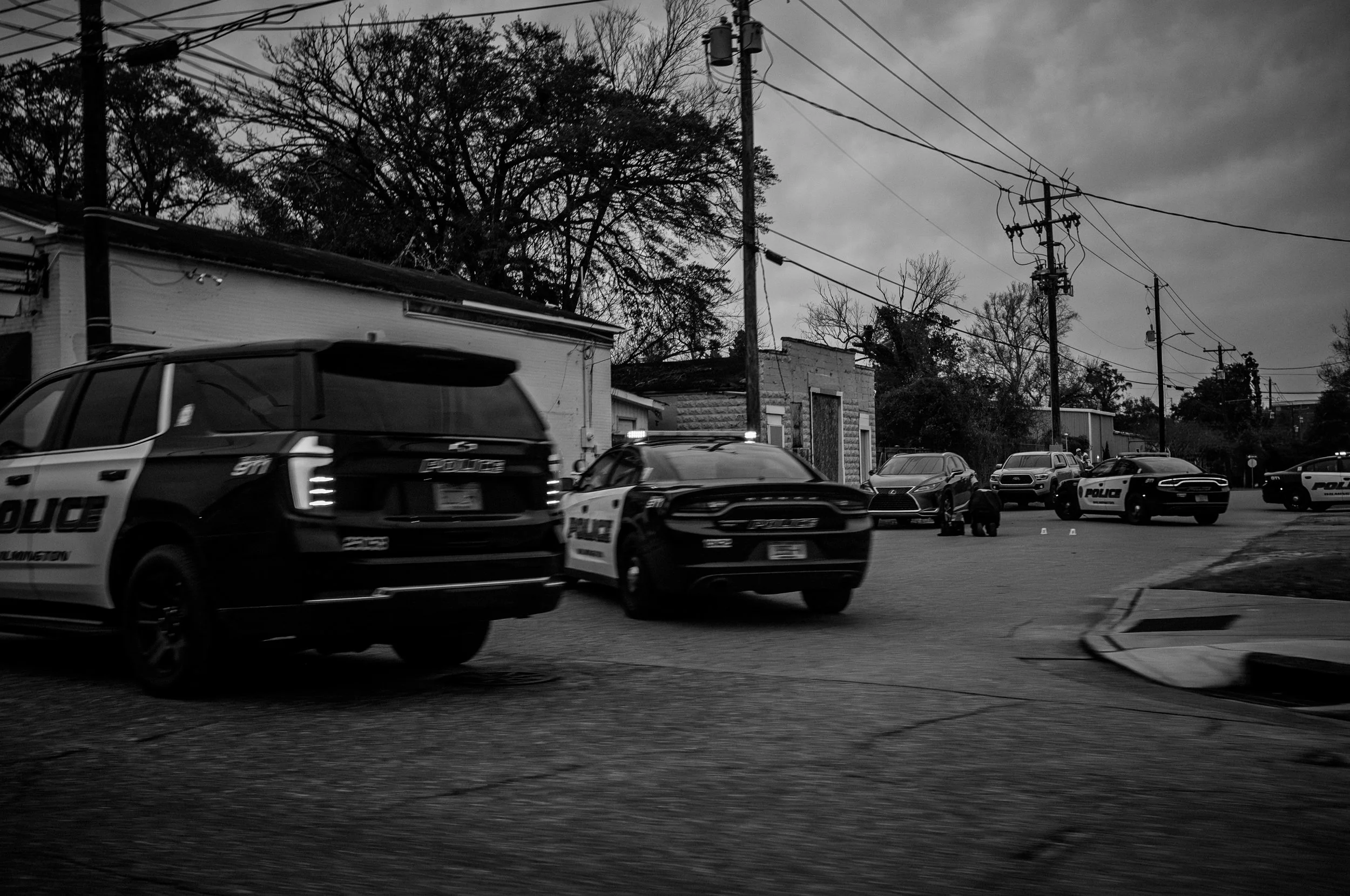 Scene of multiple police cars parked on a street in a neighborhood on a cloudy day, with some buildings, trees, and utility poles visible.