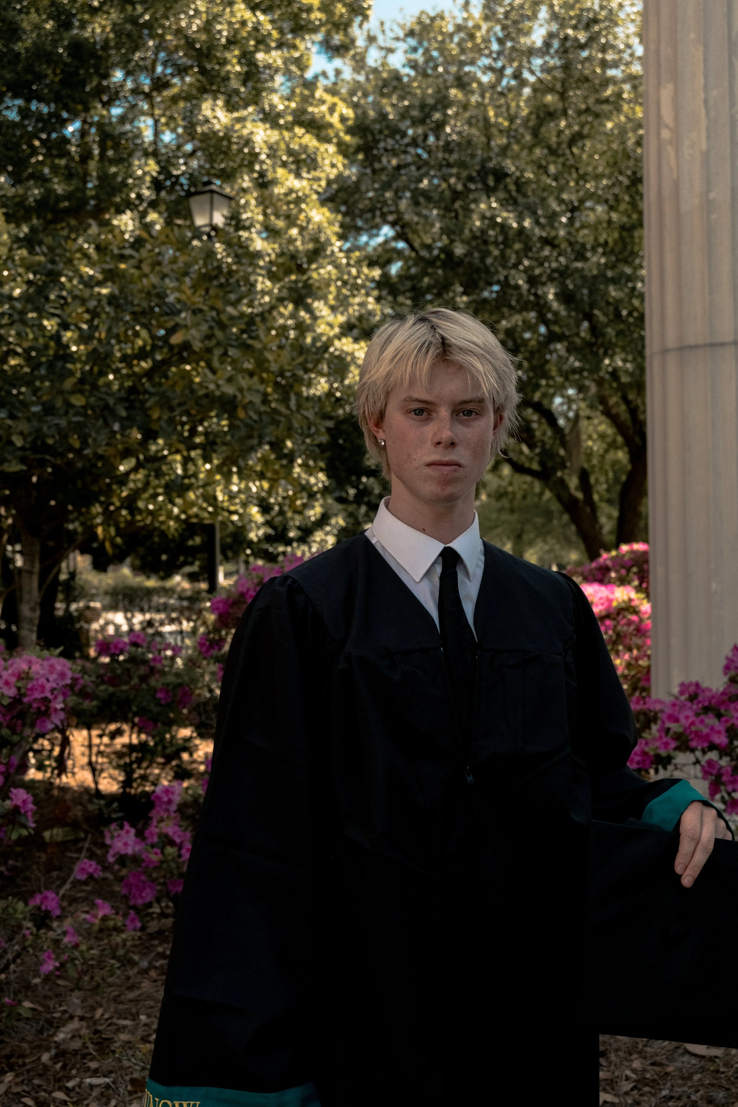 A person with short blonde hair wearing a black graduation gown and a white shirt standing outdoors near trees and pink flowers.