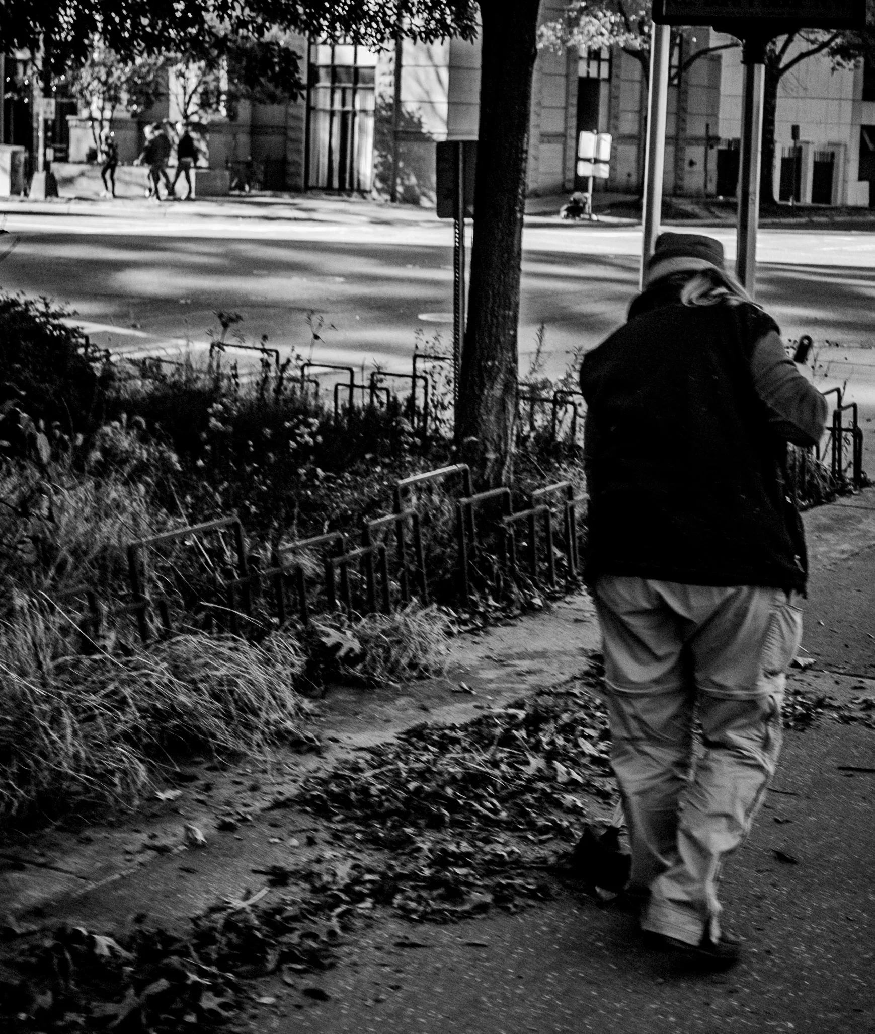 A person walking on a city sidewalk during daytime, with a street, some pedestrians, and buildings in the background.