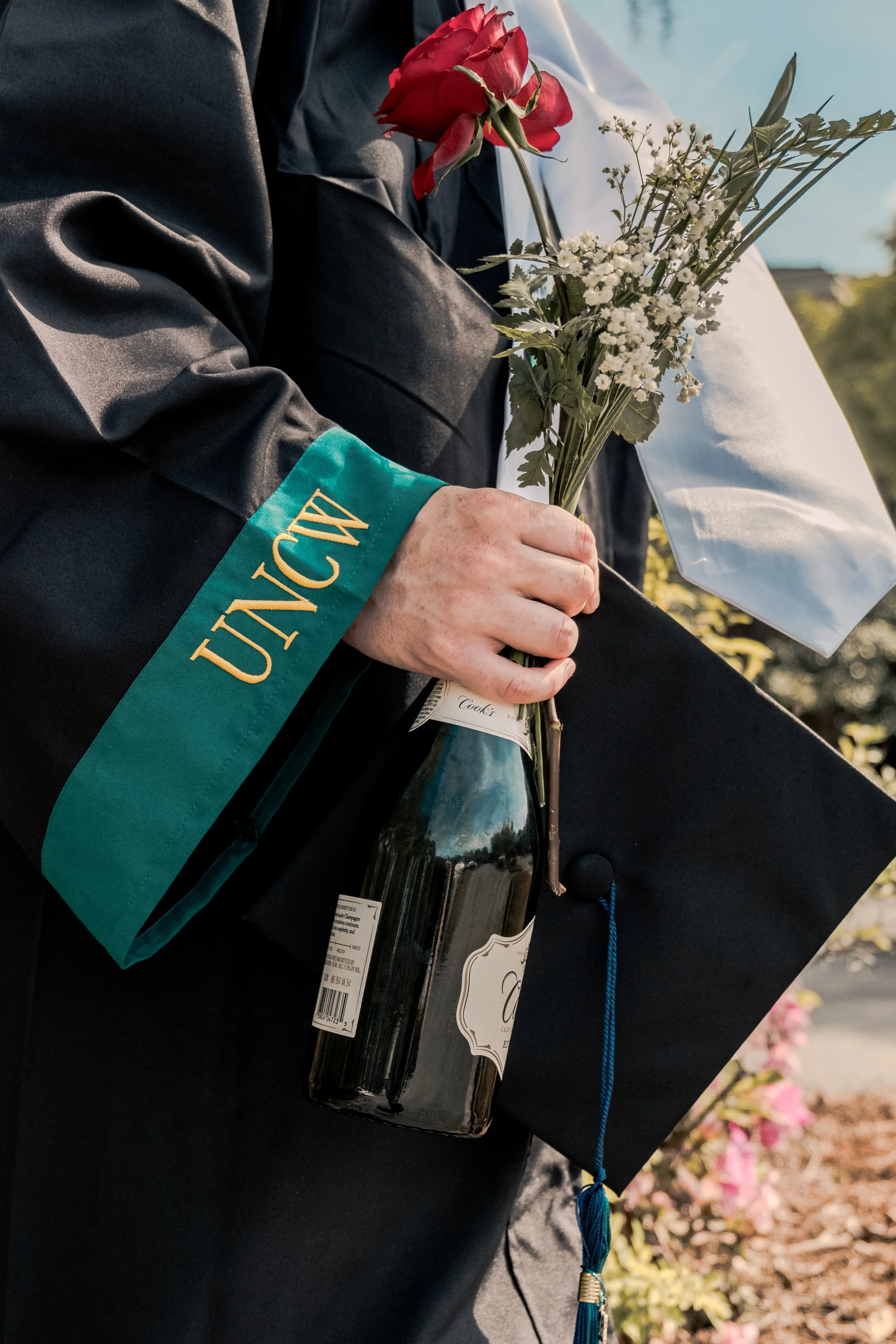 Person wearing a graduation gown holding a bouquet of flowers and a bottle of wine, with a diploma in the background.