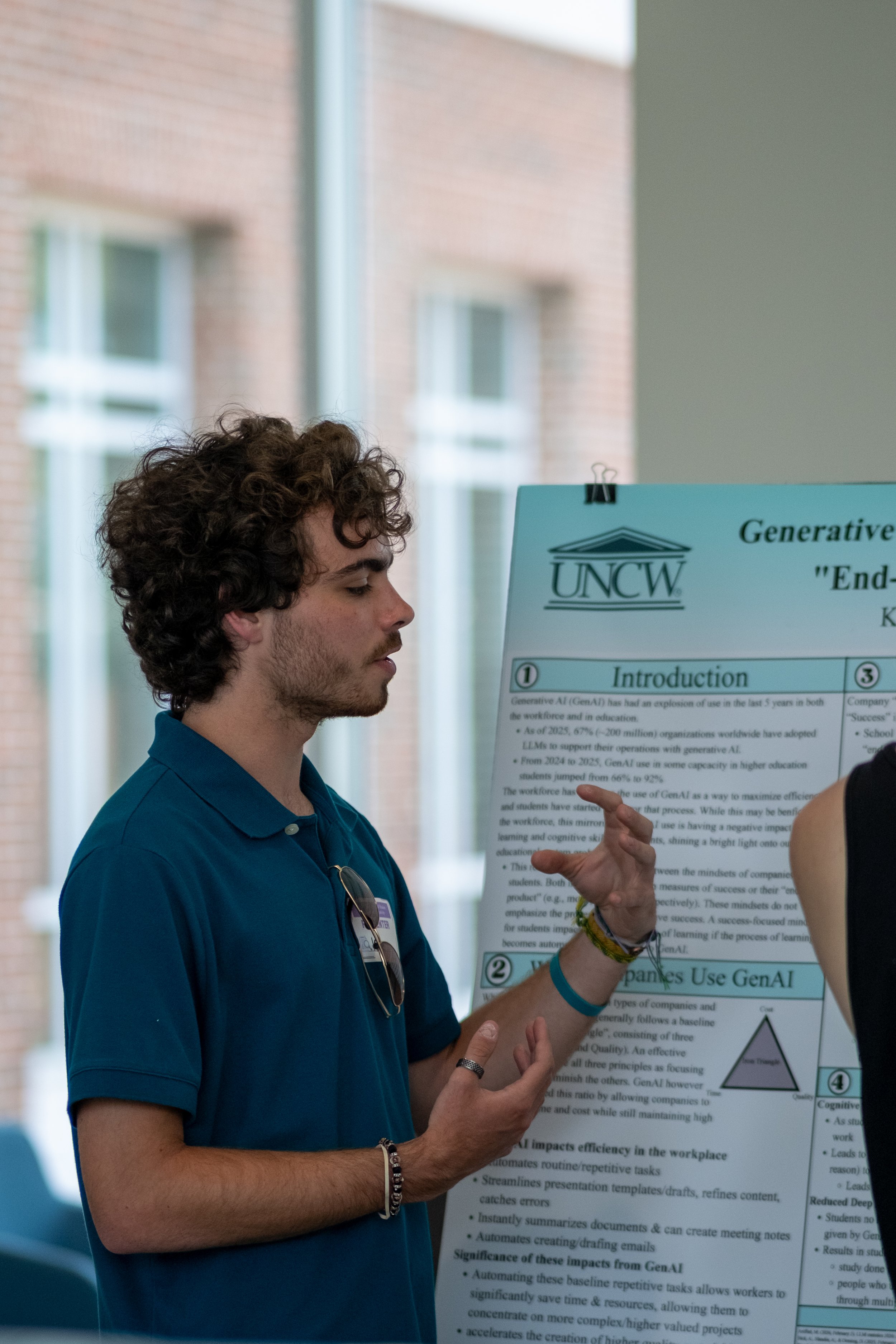 A man with curly hair wearing a blue shirt and glasses hanging from his shirt collar is presenting a research poster at an academic conference, with a background of windows and a brick building.