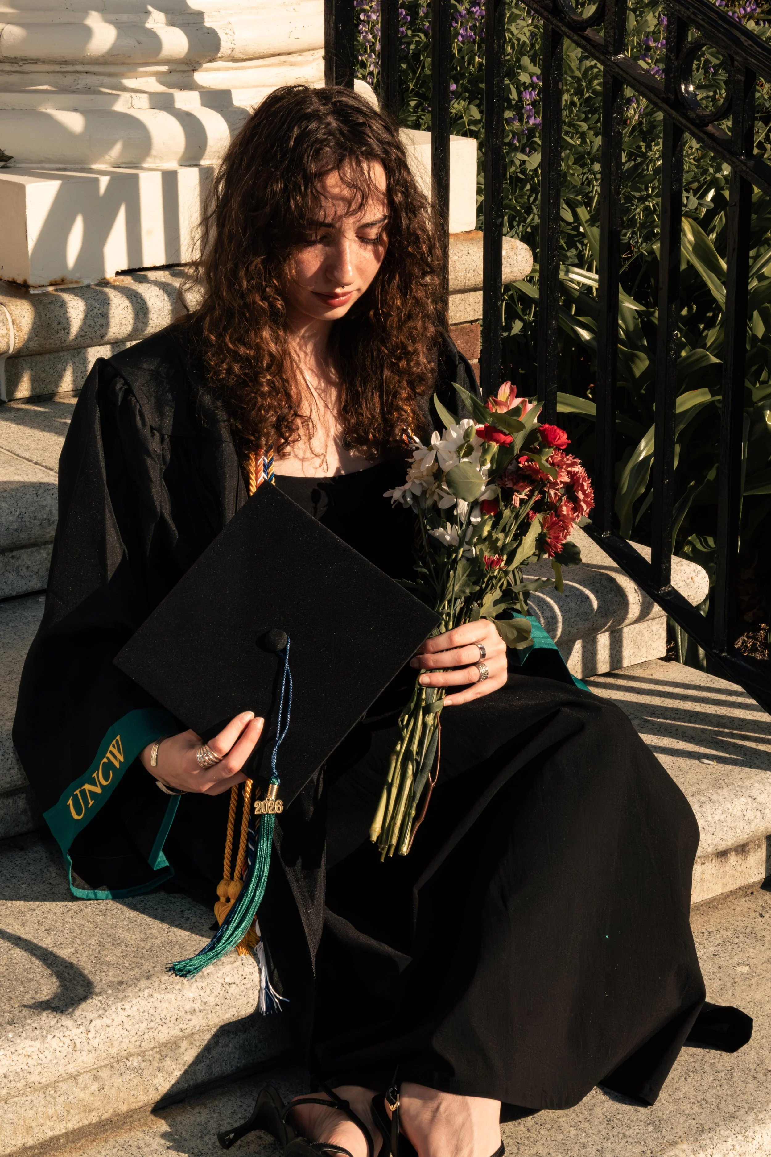 A young woman with curly hair is sitting on stone steps, wearing a graduation gown and cap. She is holding a bouquet of flowers and some graduation cords, with her eyes closed and head slightly bowed. The sunlight is casting shadows, and there is a b