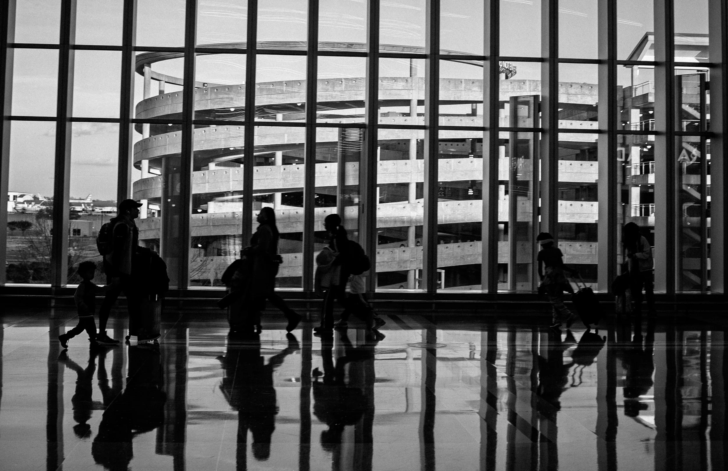 Silhouettes of people walking inside an airport terminal with large windows and a parking garage visible outside.