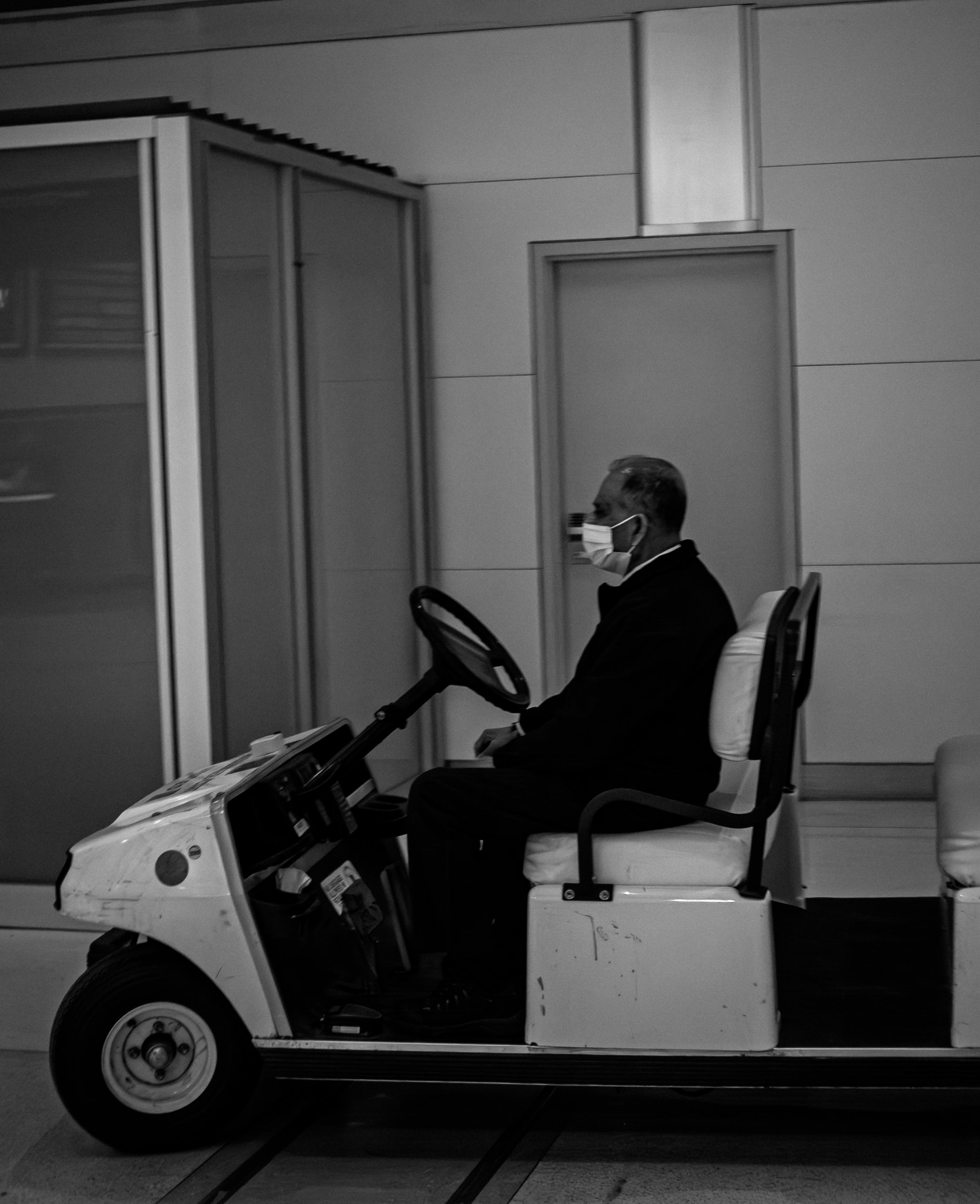 A man wearing glasses and a face mask sitting on a small electric vehicle or golf cart in an indoor setting.