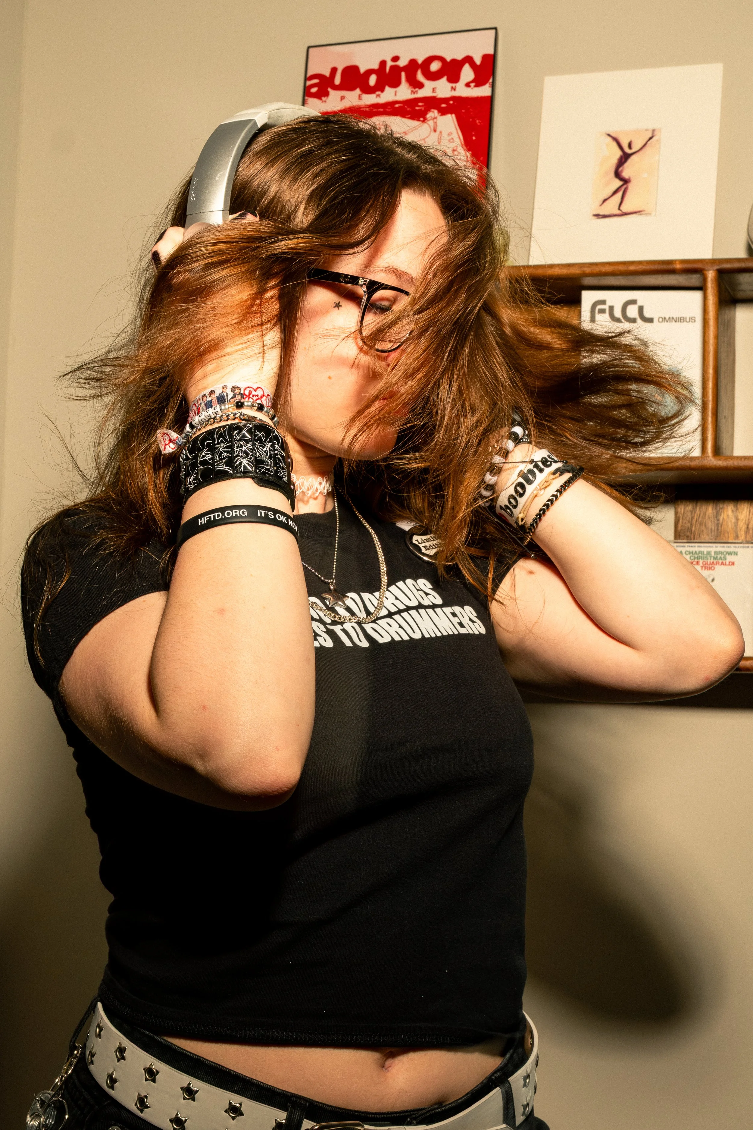 A young woman with long brown hair wearing headphones, glasses, multiple bracelets, a black t-shirt with white text, and a white belt with star-shaped studs. She is adjusting her headphones with her eyes closed and hair falling across her face.