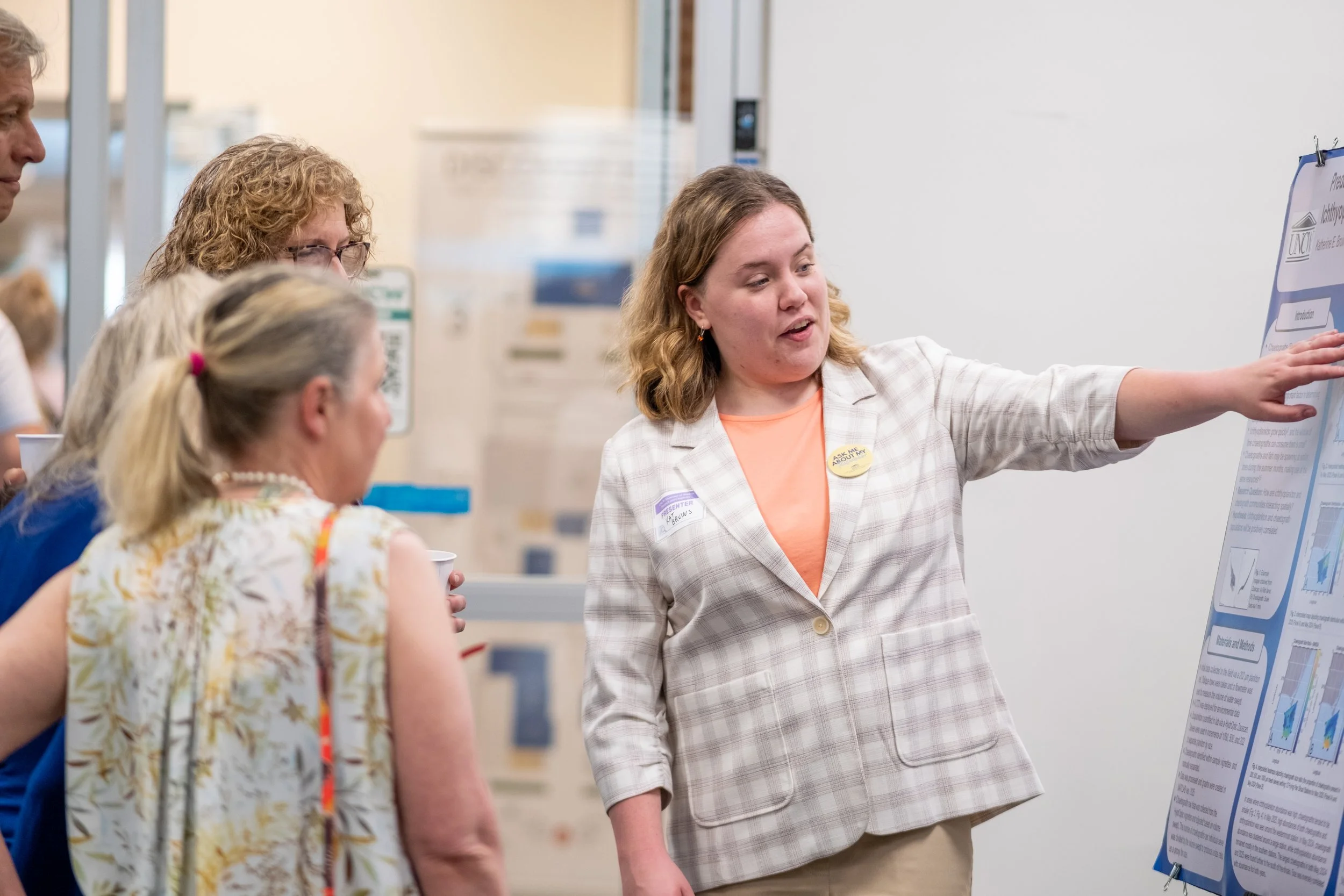 A woman with shoulder-length wavy hair wearing a beige checkered blazer and an orange shirt is giving a presentation at a science or academic conference. She is pointing towards a large poster with text and images taped to a whiteboard. Four people a