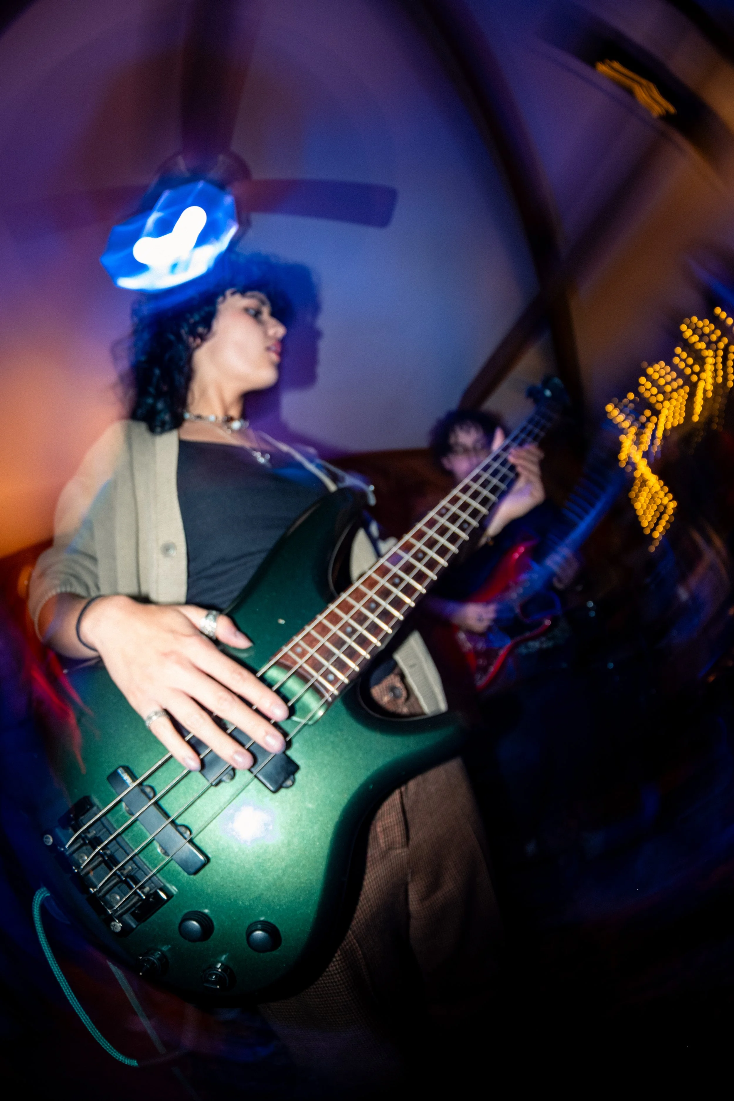 A woman with curly dark hair playing a green electric bass guitar in a dimly lit room with purple lighting.