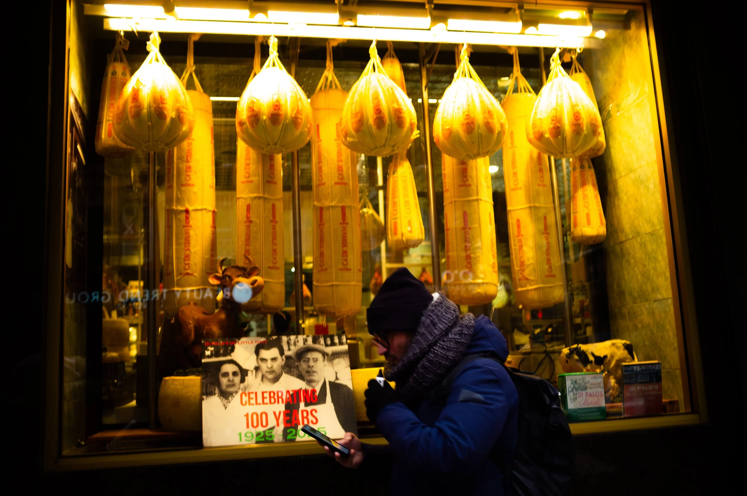 A person wearing a dark winter coat, beanie, and scarf looking at a smartphone outside a storefront window. The window displays hanging cured meats, a cow figurine, and a sign celebrating 100 years from 1925 to 2025.