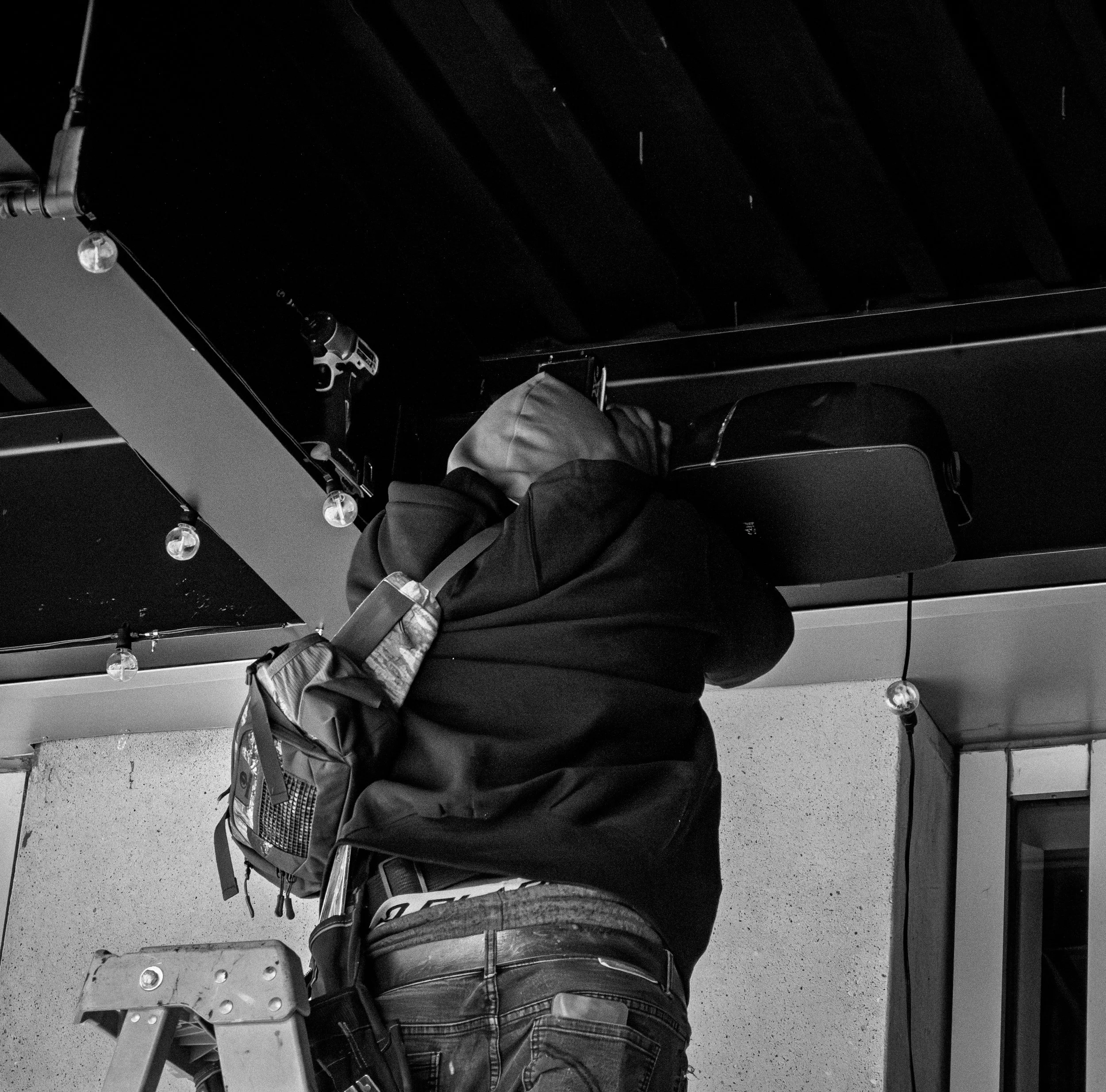 Person working on a black table with a drill and various tools, wearing a backpack, in an indoor space with string lights.