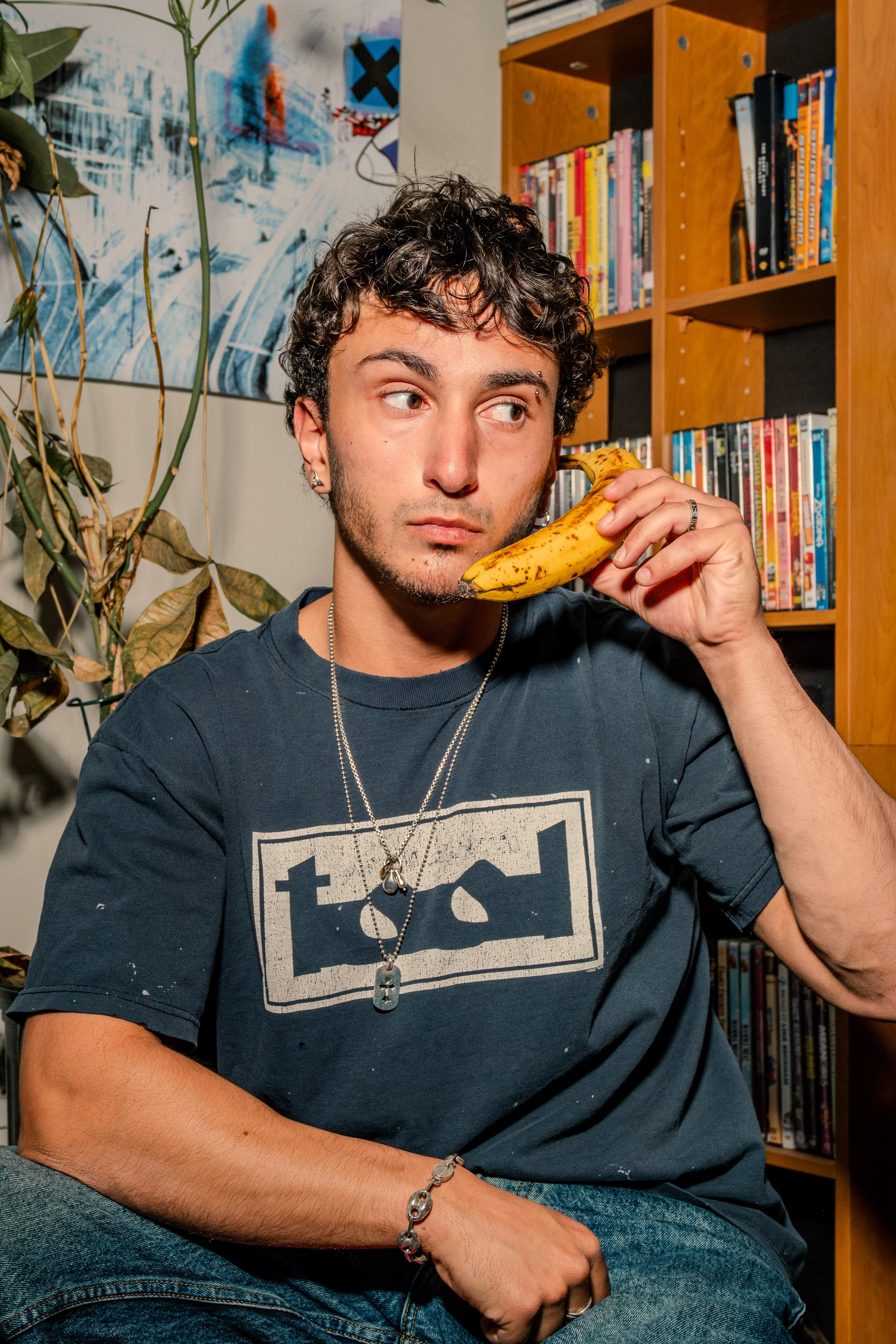A young man with curly dark hair and earrings holds a ripe banana to his ear as if it were a phone, seated indoors beside a bookshelf and a plant.