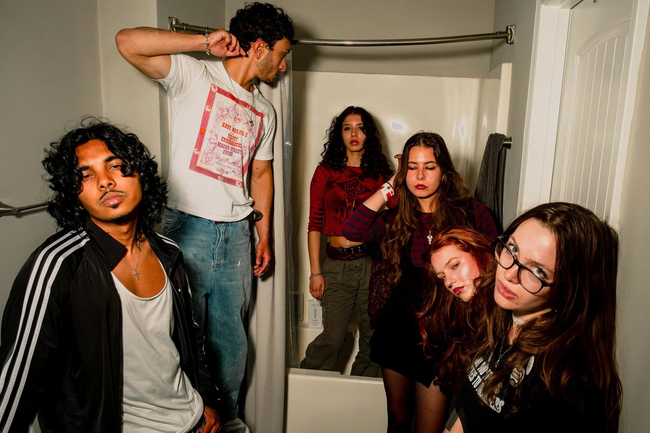 A group of young people with diverse appearances in a small bathroom, some looking into a mirror and others posing for a photo.
