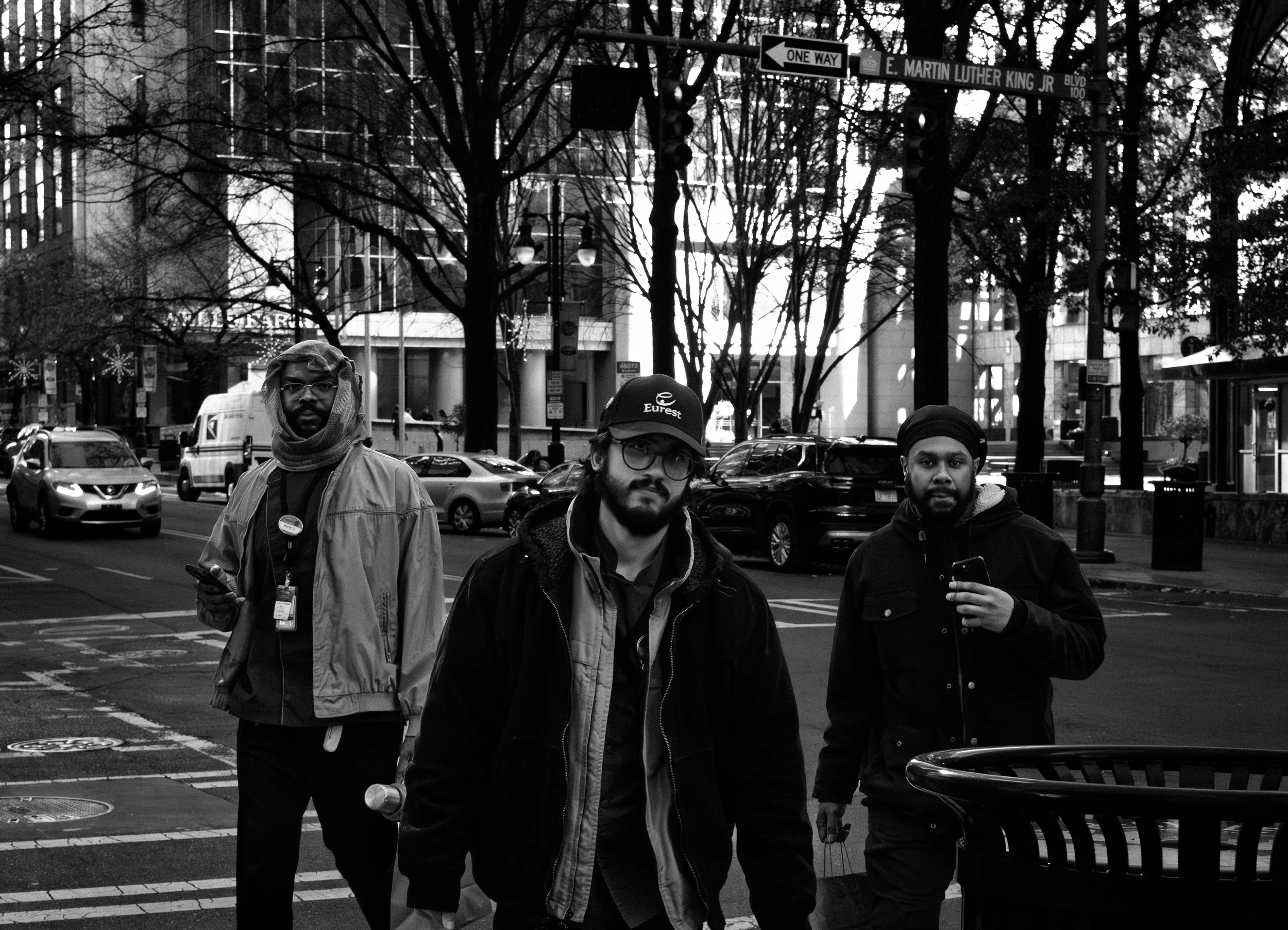 Three men walking on a city street in black and white, with trees, cars, and tall buildings in the background.