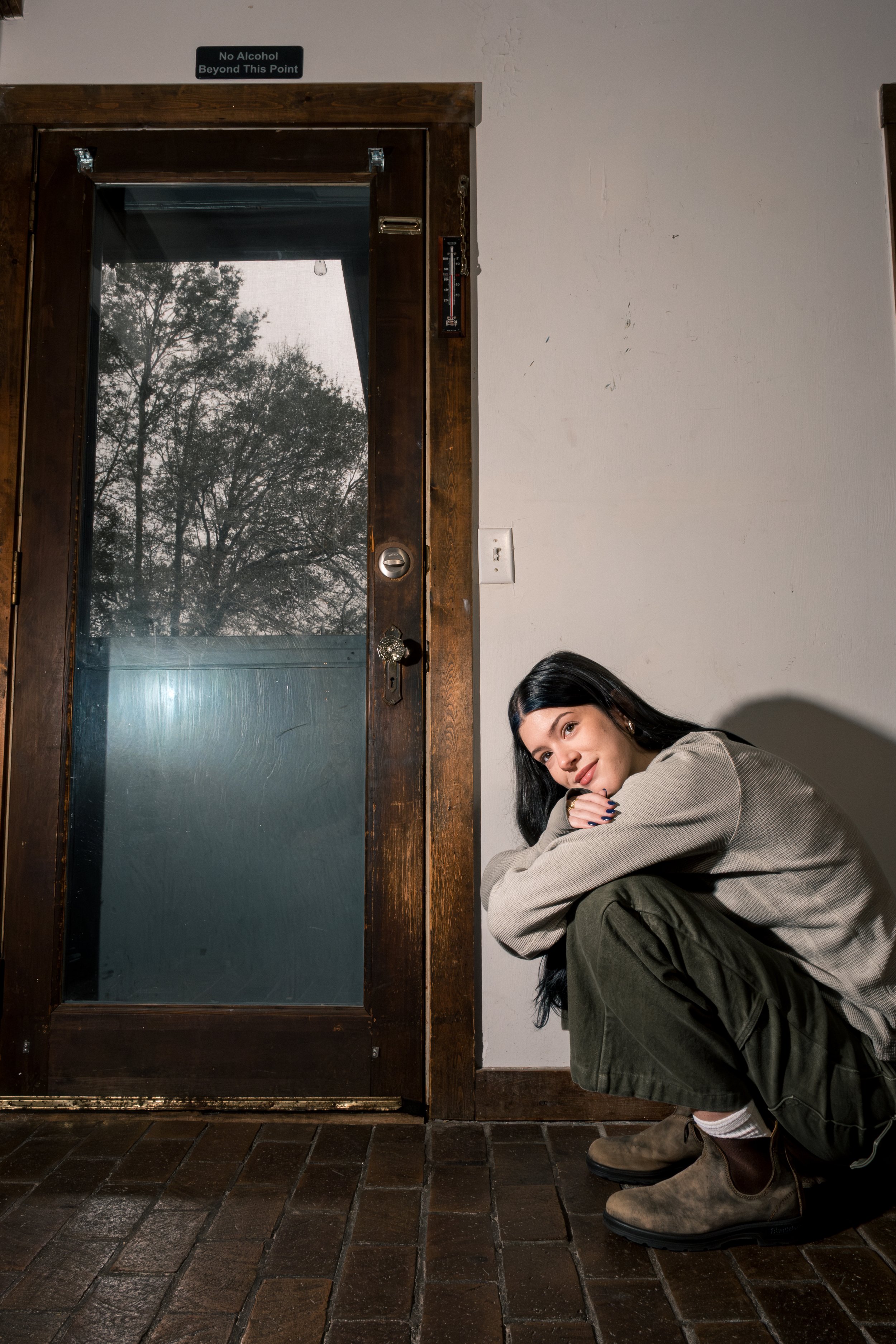 A young woman with long dark hair crouching near a wooden door with frosted glass. She is wearing a beige jacket, olive green pants, and brown boots, smiling at the camera. A sign above the door reads, 'No Alcohol Beyond This Point,' and there is a t
