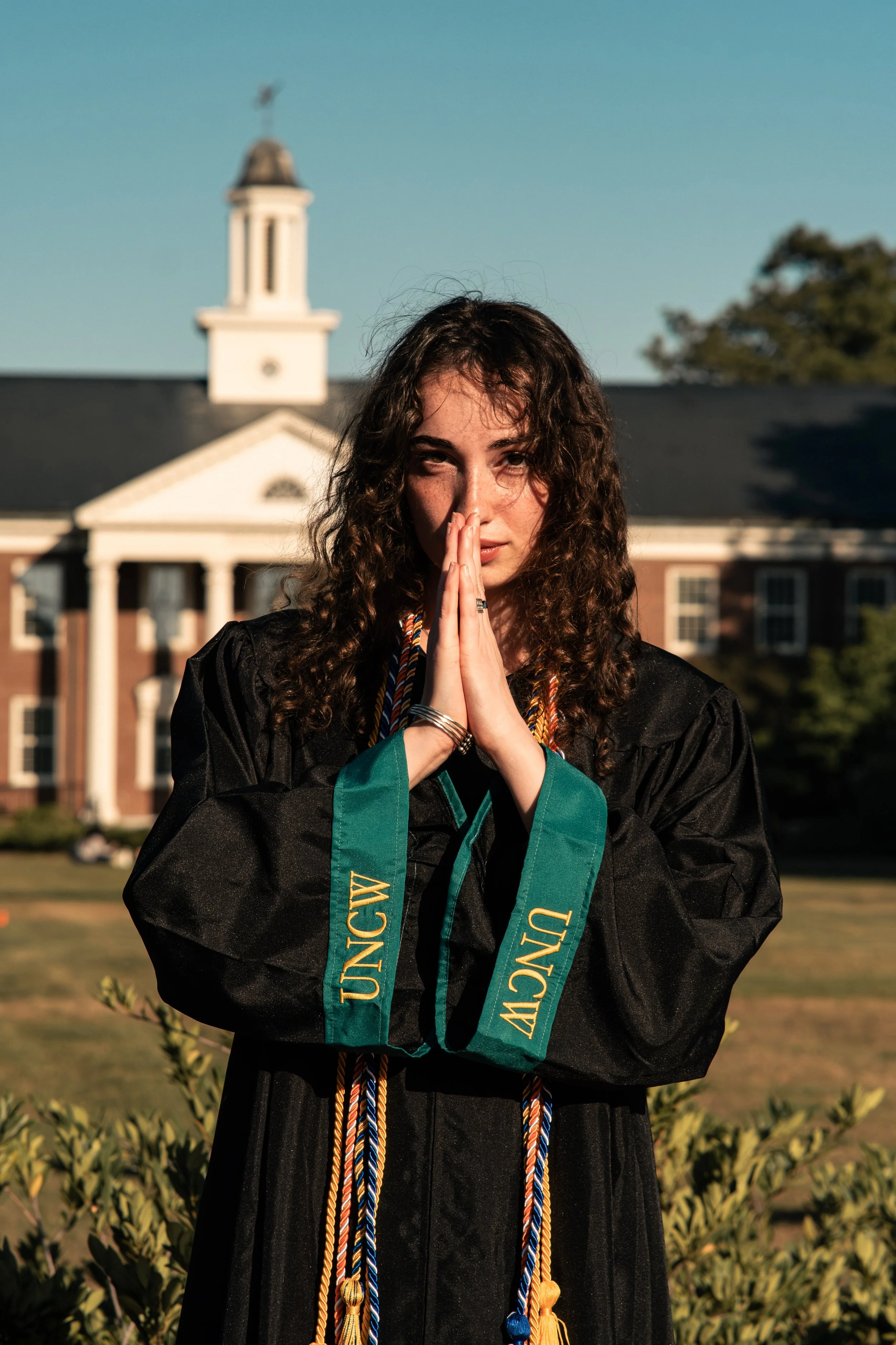 A young woman with curly hair in graduation gown and cords, standing outdoors in front of a college building, with her hands pressed together near her face.