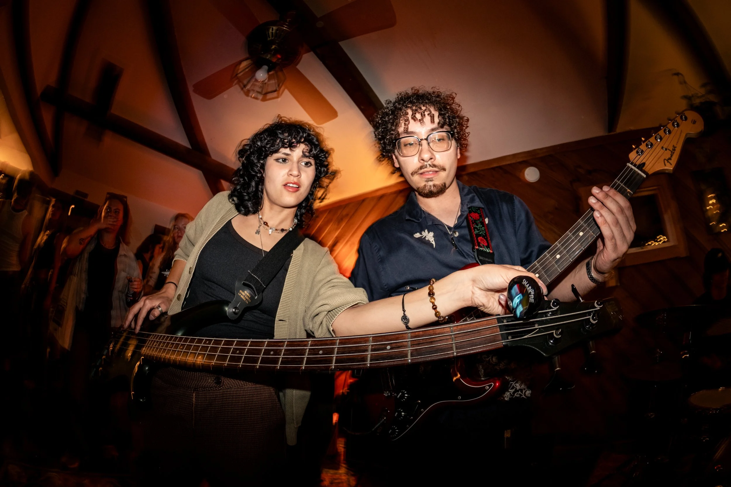 Two musicians, a woman and a man, playing guitar together indoors with dim lighting. The woman has curly black hair and wears a black top with a beige jacket, while the man has curly hair, glasses, and a dark shirt with floral embroidery. There are o