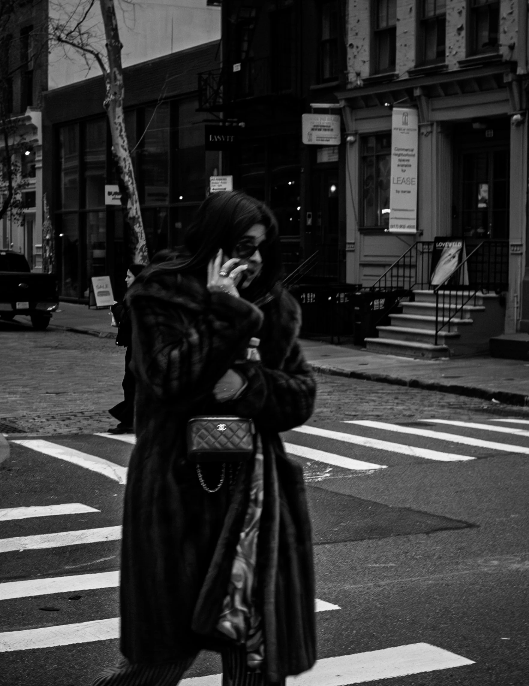 A woman in a long coat and sunglasses talking on a cell phone while crossing a city street at night. The background features storefronts, signs, and a staircase leading to a building entrance.