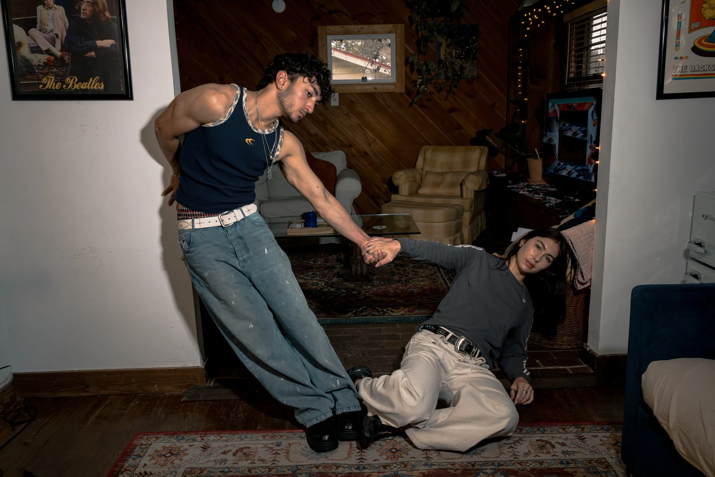 Two young adults, a man and a woman, are in a living room. The man is standing and leaning over the woman, who is lying on the floor. They are holding hands and looking at each other. The room has wood paneling on the walls, framed pictures, and a te