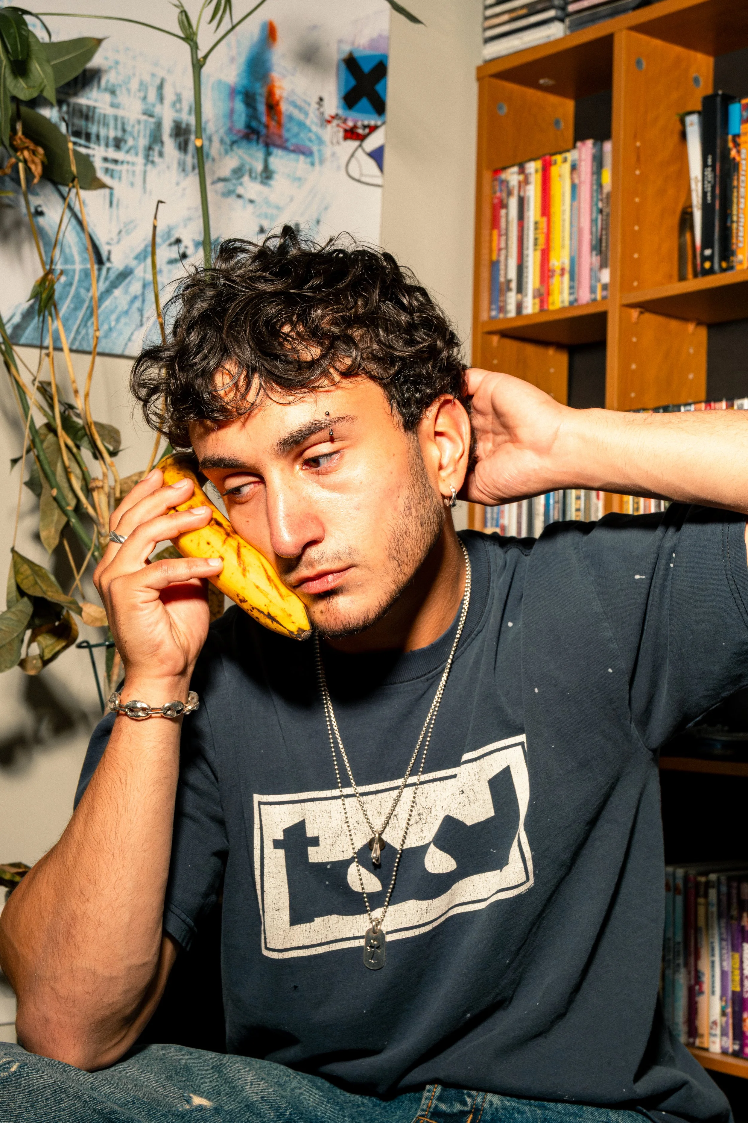 A young man with curly hair and pierced eyebrows holding a banana to his ear like a phone, sitting in front of a bookshelf and a colorful painting.
