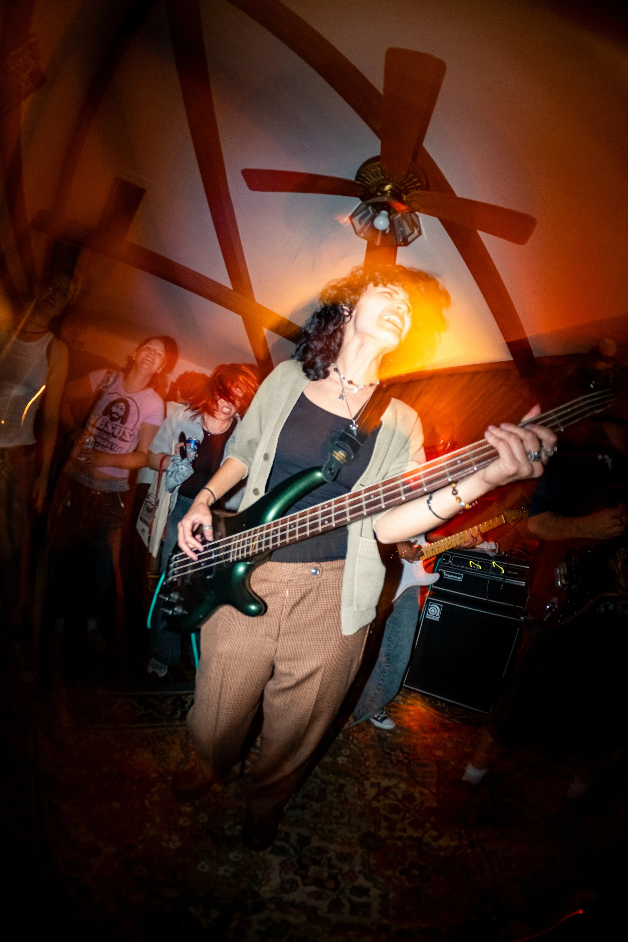 A woman with curly hair playing an electric bass guitar at a musical performance, with people in the background and a ceiling fan overhead in a cozy indoor setting.
