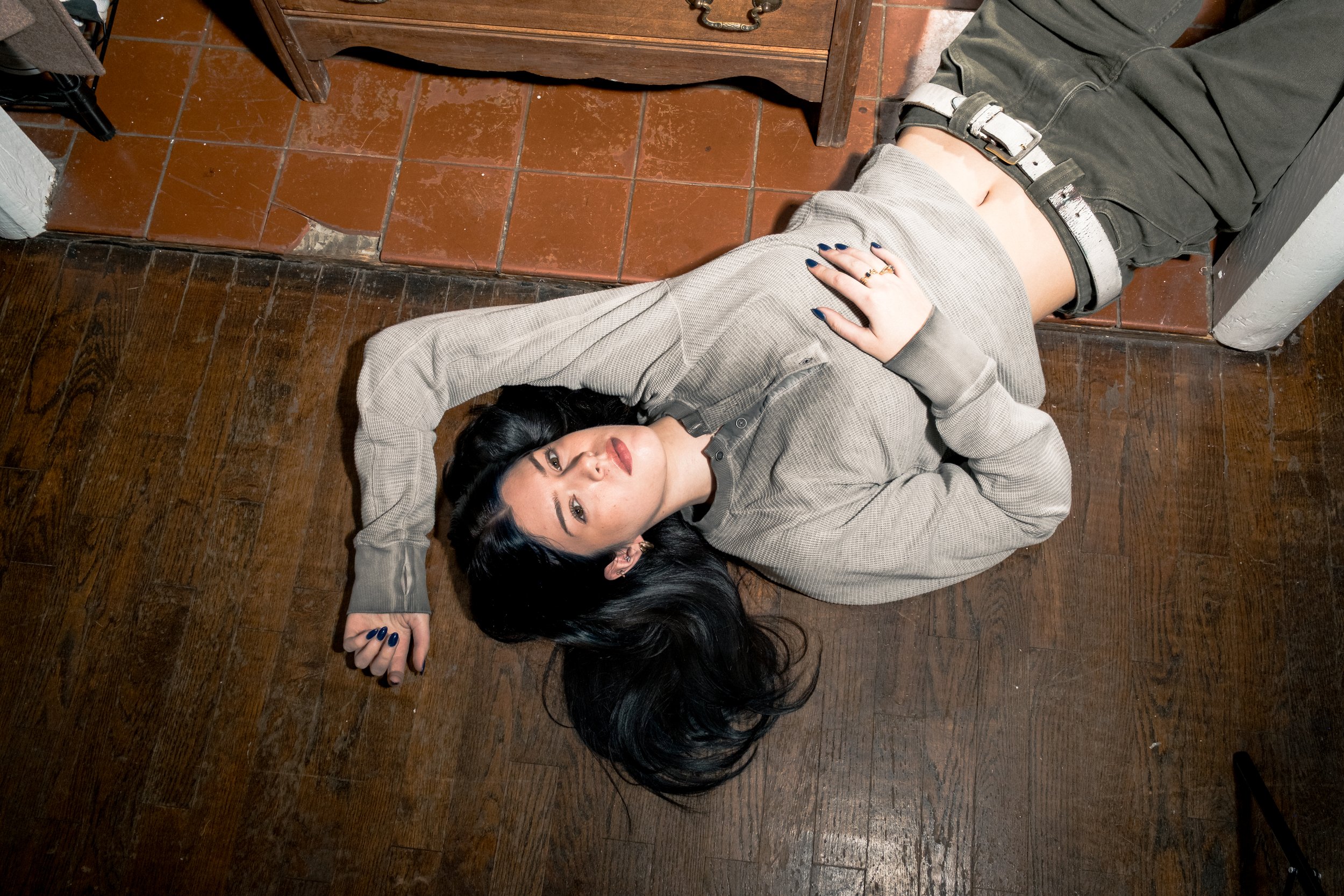 A woman with long black hair lying on the wooden floor with her head near a woman with dark hair, who is lying on a tiled floor. The woman on the wooden floor is looking up at the camera, wearing a light-colored jacket. The woman on the tiled floor i