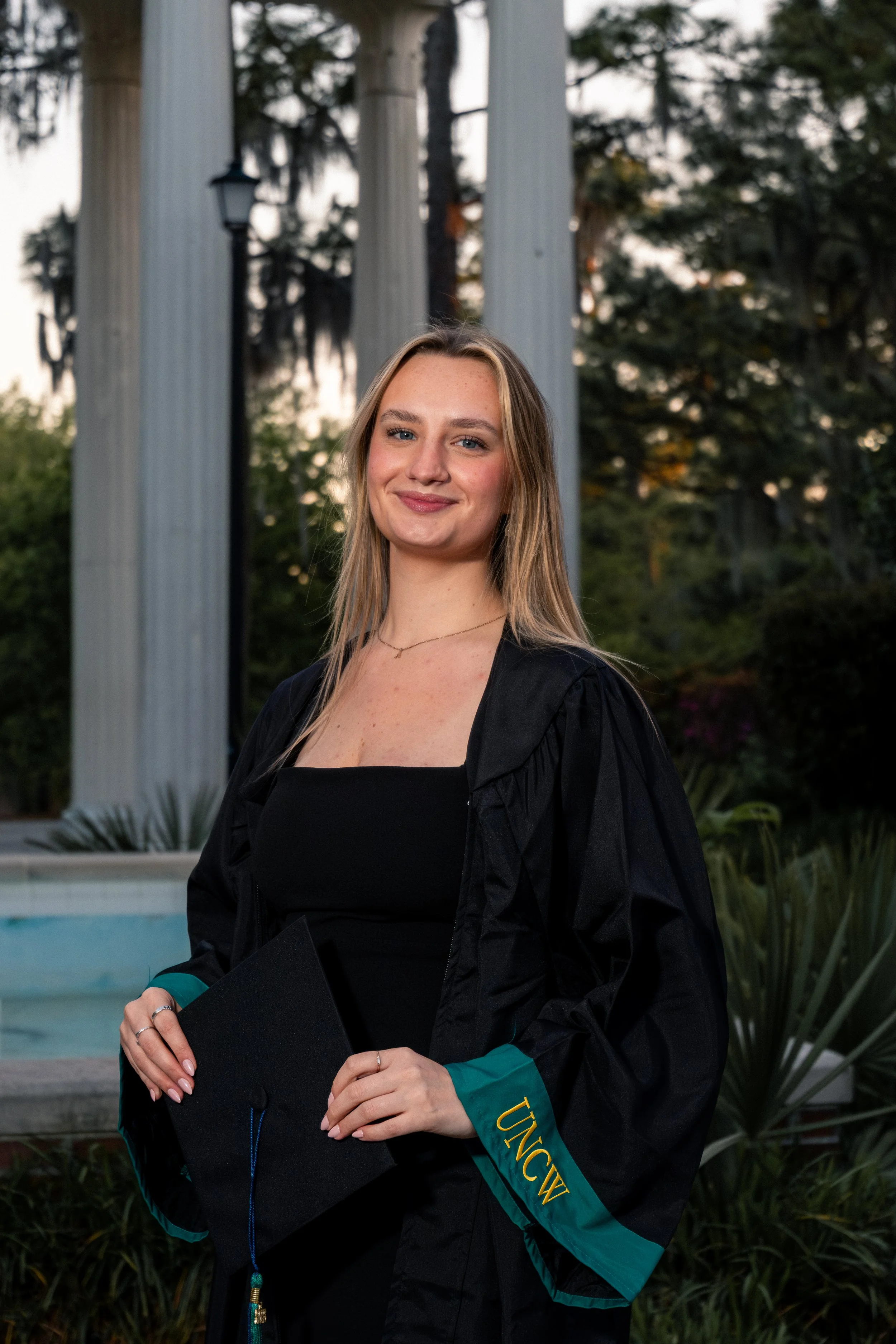 A young woman in a black graduation gown with teal accents, holding a diploma, standing outdoors near a pool with trees and large white columns in the background.
