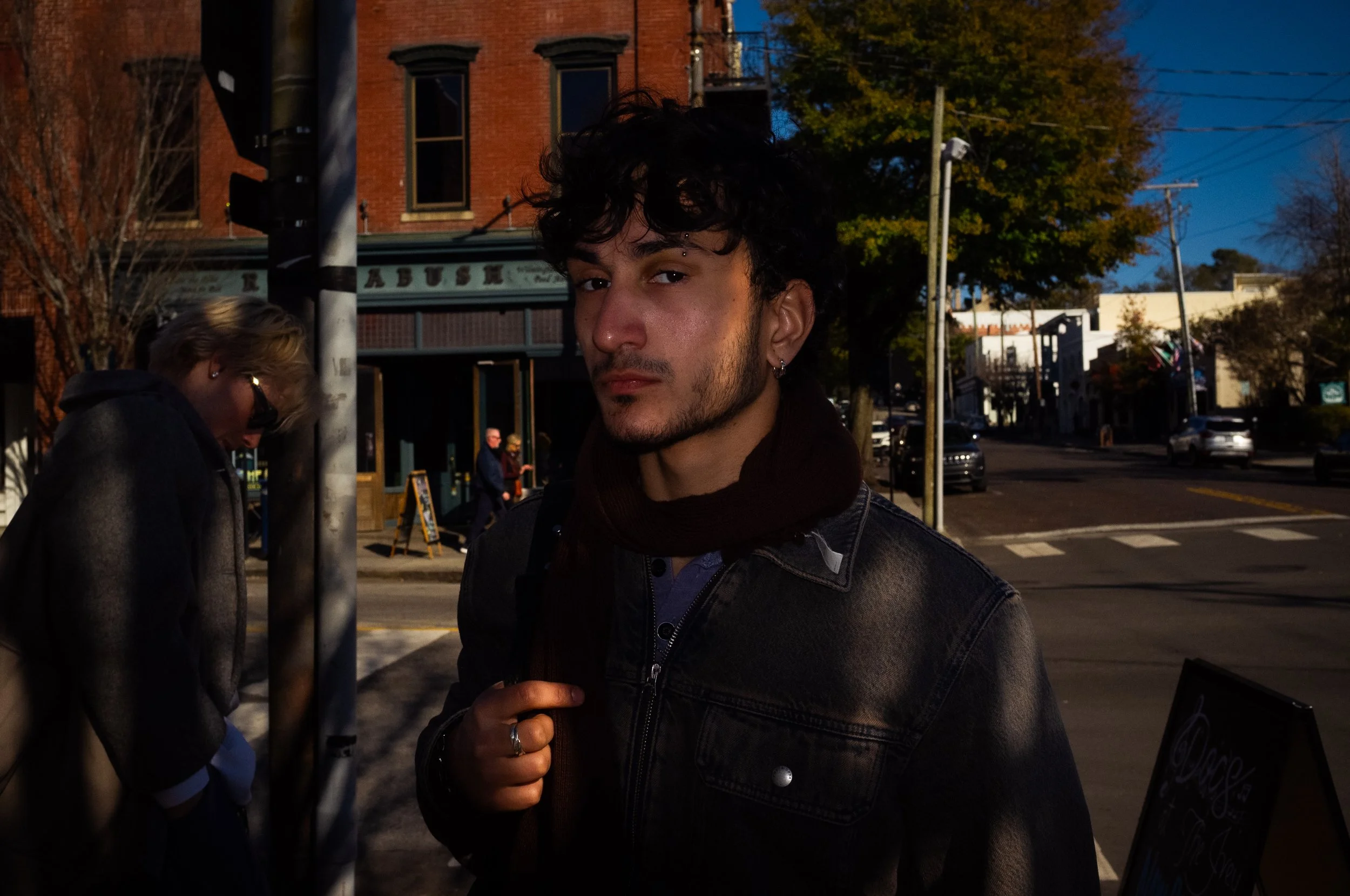 A young man with curly dark hair and earrings standing on a city street at dusk, with a brick building, trees, and cars in the background.