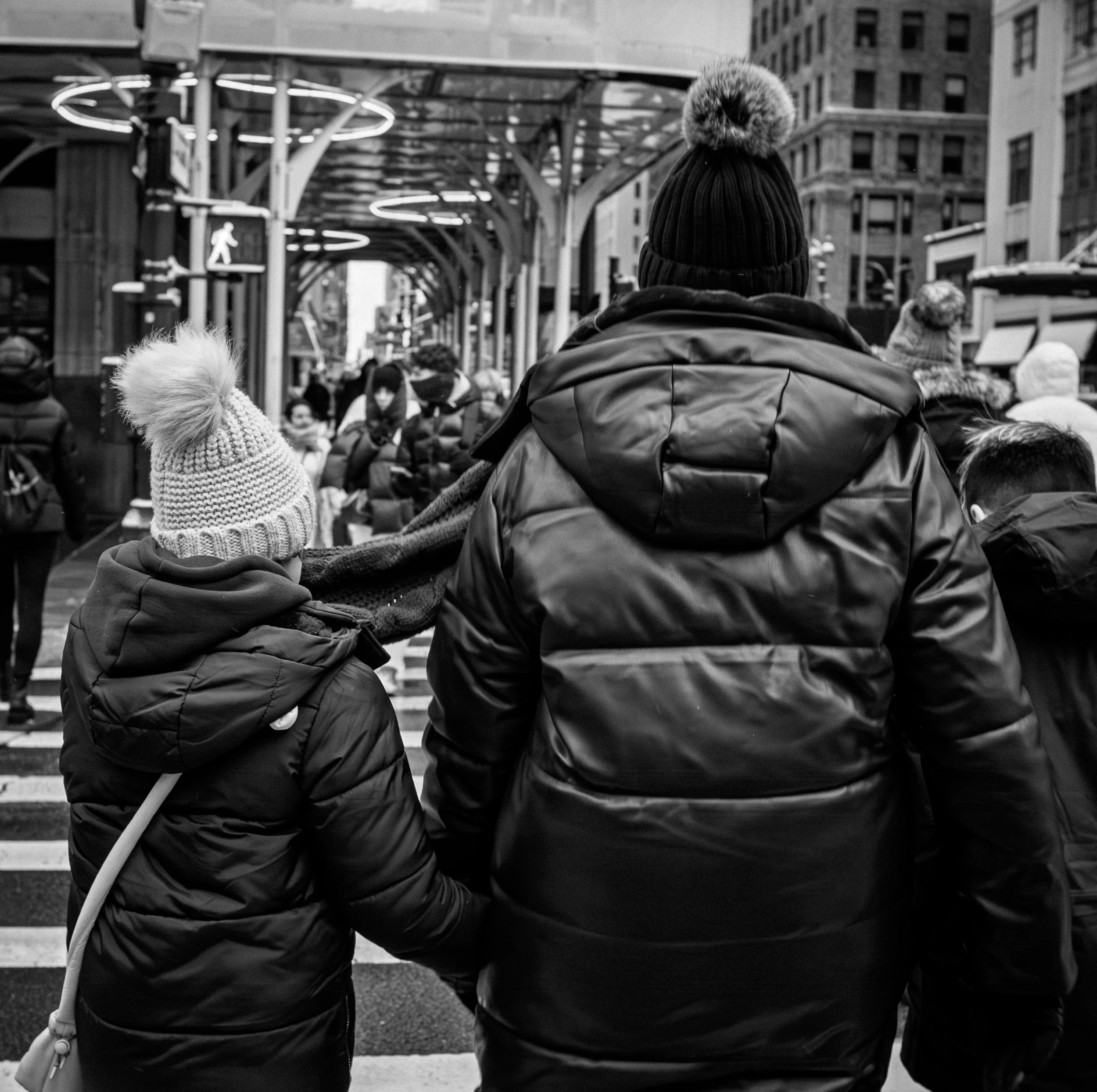 People crossing a city street, wearing winter clothing; a child in a knitted hat with a pom-pom, an adult in a beanie and a puffy jacket, other pedestrians in the background.