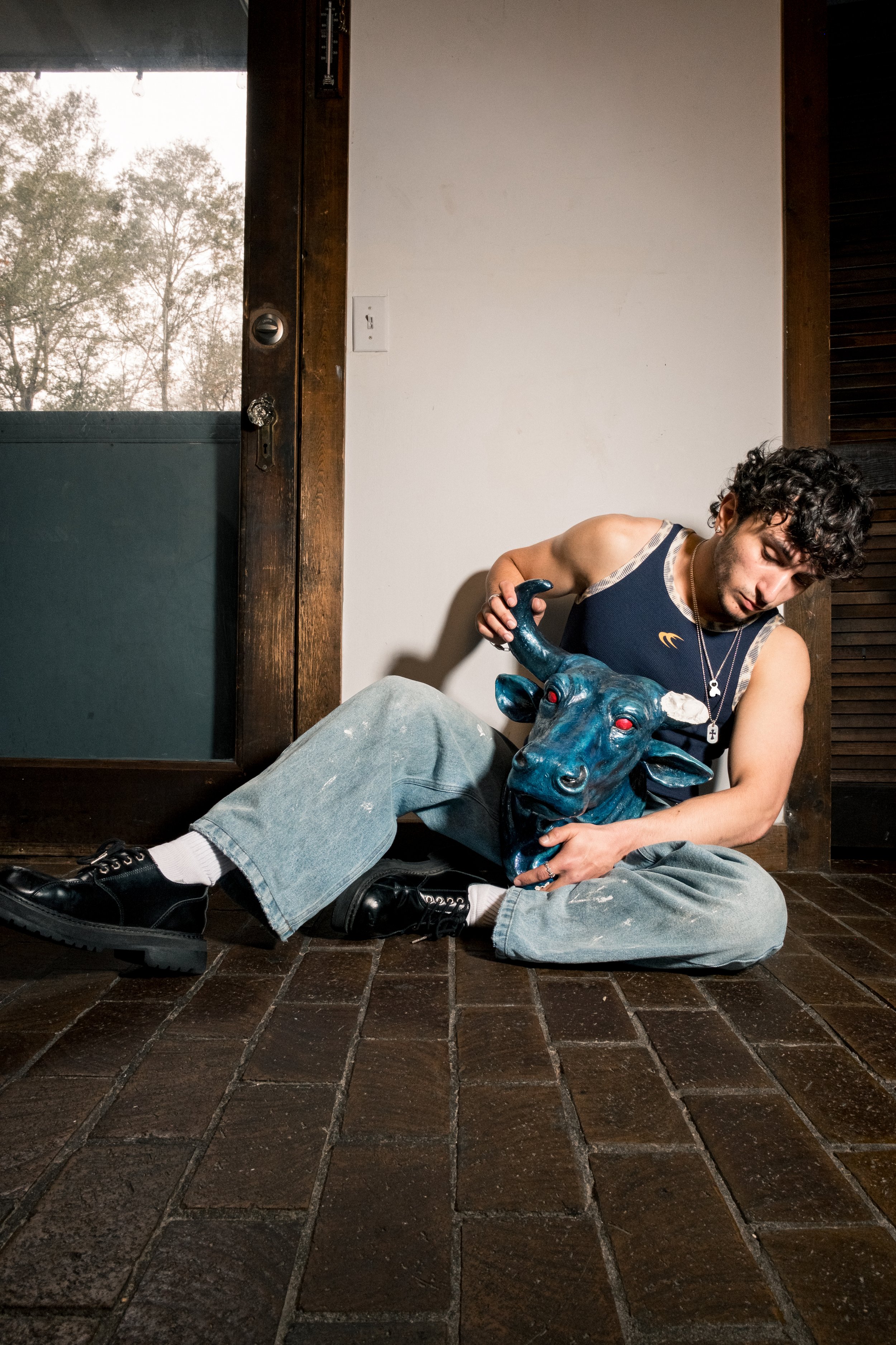 A young man with curly dark hair sitting on a dark tiled floor, wearing a sleeveless navy shirt, distressed jeans, and black shoes. He is holding a blue bull sculpture with red eyes and appears to be examining or resting on it. The background include
