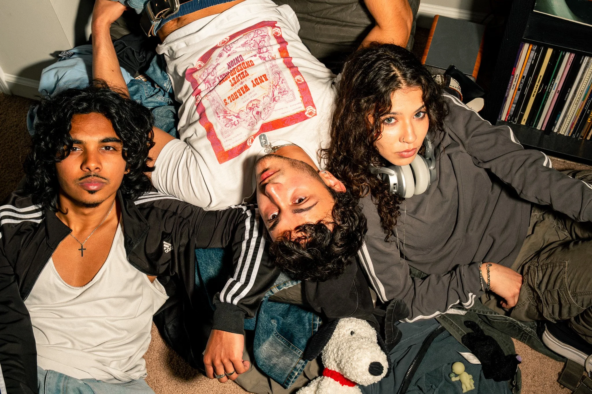 A group of four young people lying on the floor, holding hands. The person in the center has curly brown hair, wearing headphones around their neck, and is looking at the camera. The person on the left has dark wavy hair, a cross necklace, and is wea