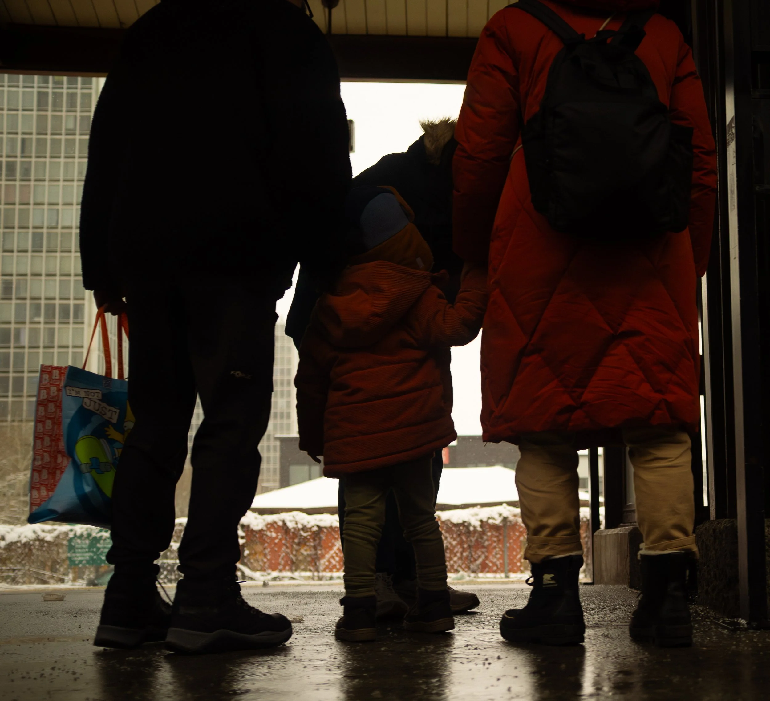 A family standing together at an entrance, holding hands, with snow outside and tall buildings in the background.